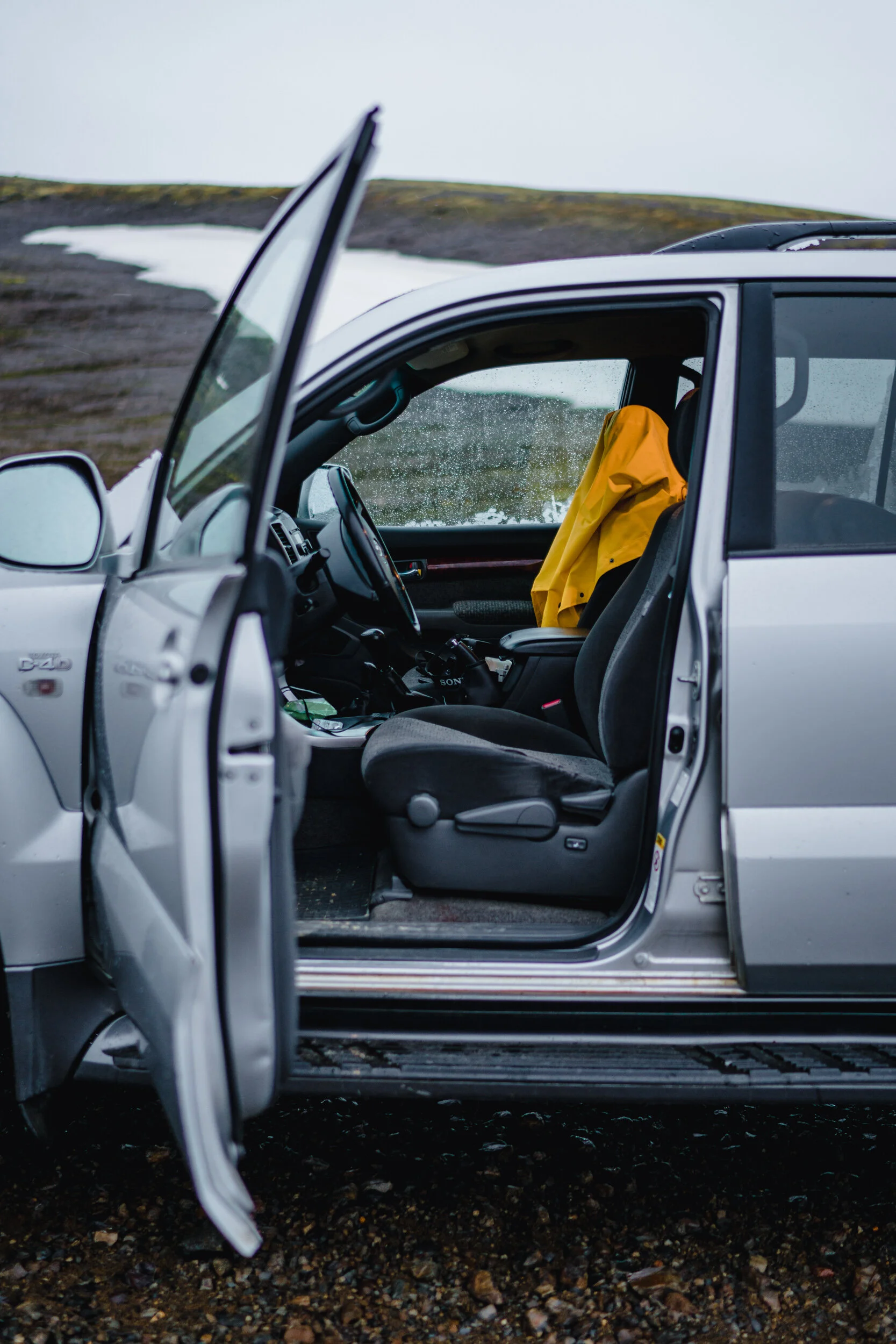 A silver car with an open door parked on a rocky ground near a snow-covered hillside, rain on the windows, and a person wearing a yellow jacket sitting inside.