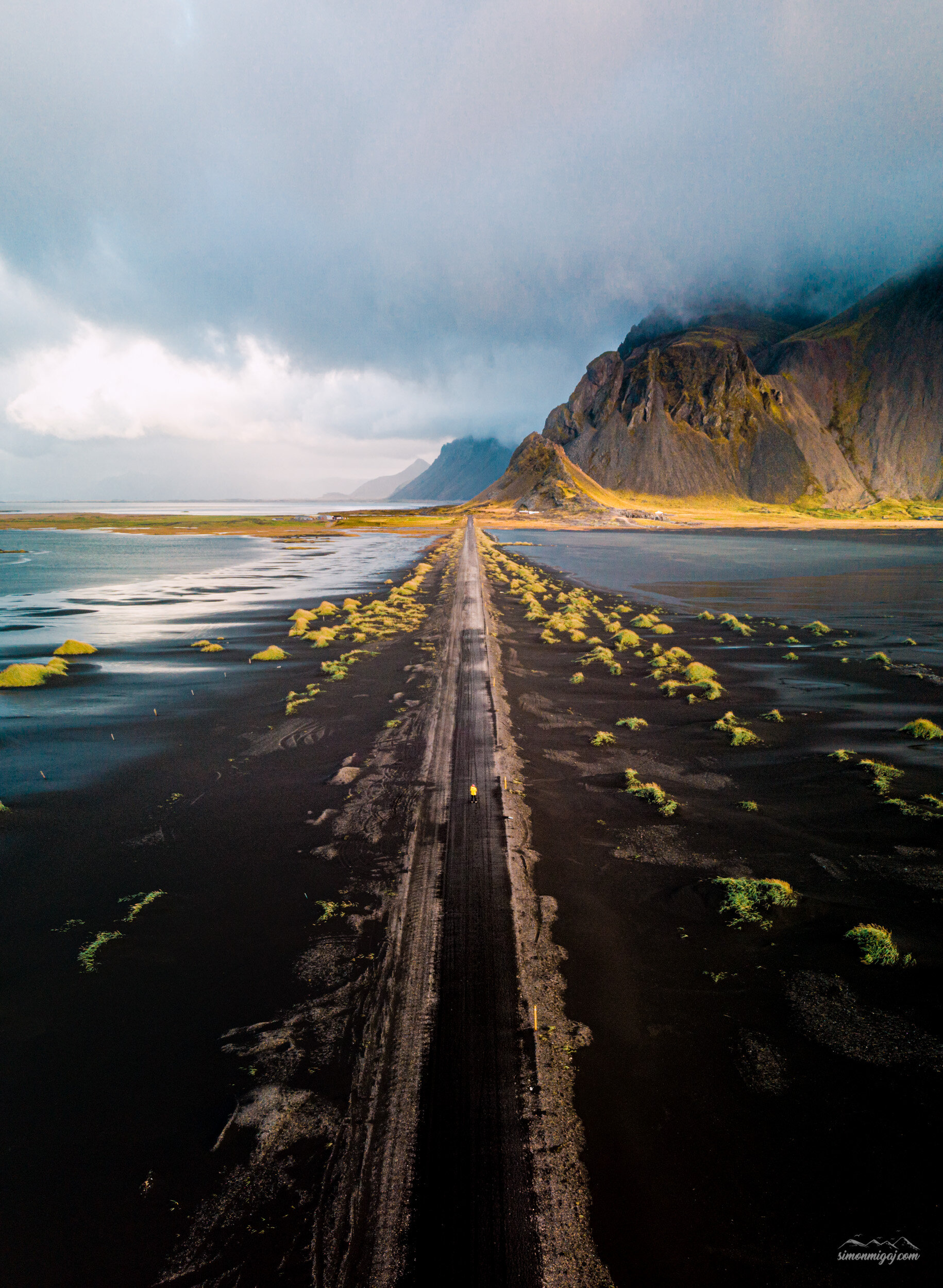 A long, straight dirt road running through a dark, marshy landscape, leading toward snow-capped mountains shrouded in clouds in the distance. The sky is cloudy and overcast.