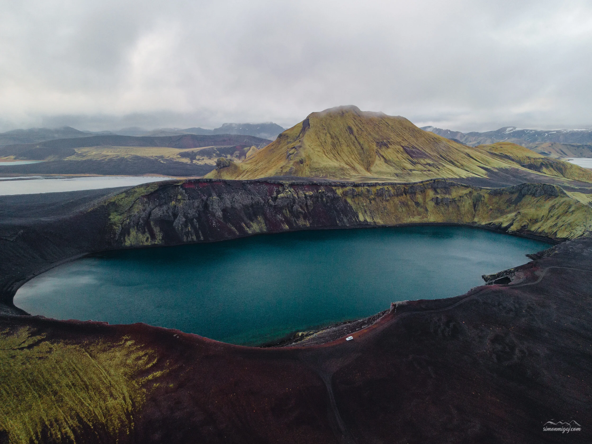 A scenic view of a volcanic crater lake with dark volcanic terrain and green moss-covered slopes, surrounded by mountains under a cloudy sky.