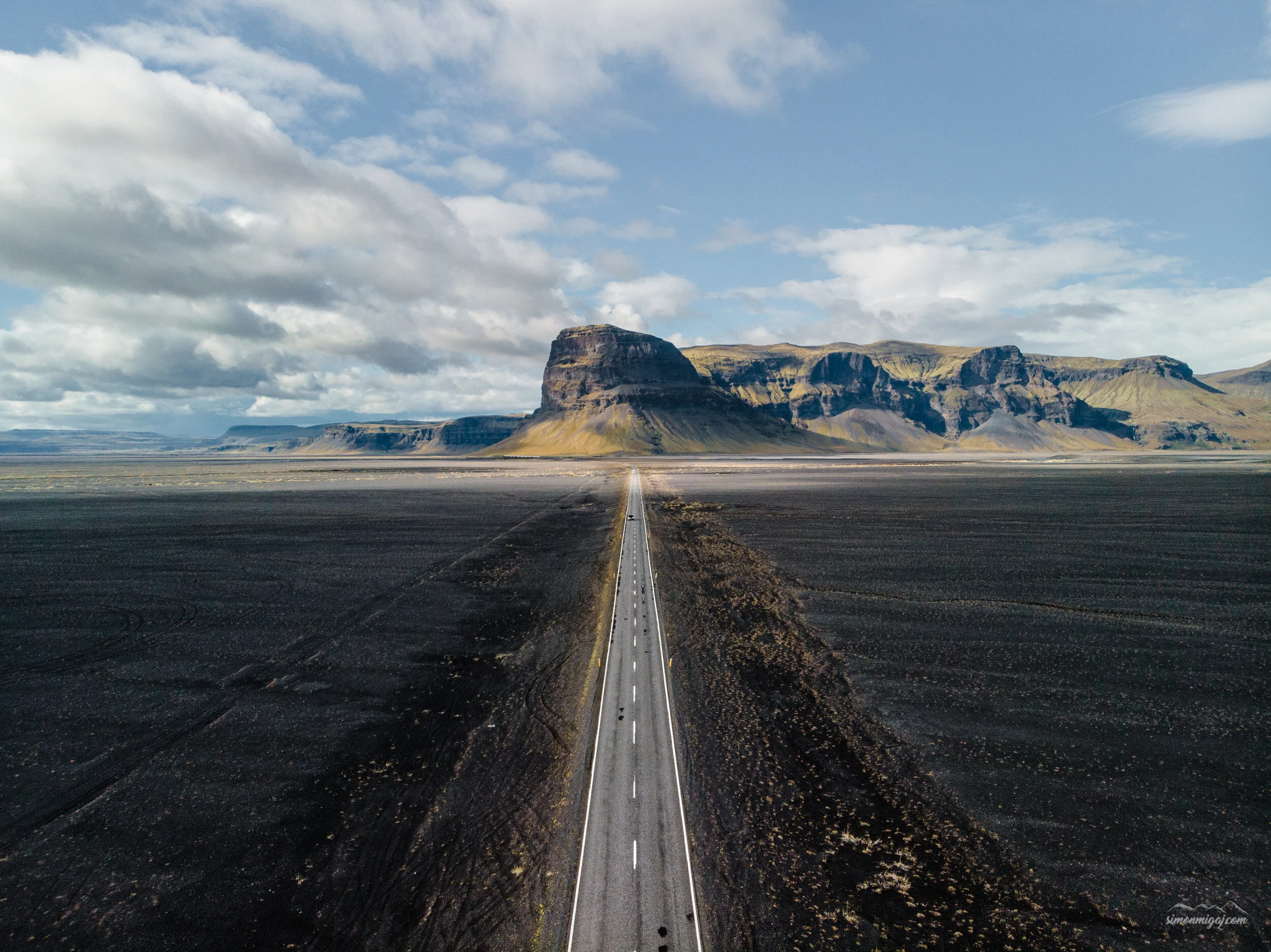 A long straight road leading towards a large, flat-topped mountain in a vast, open landscape with cloudy sky.