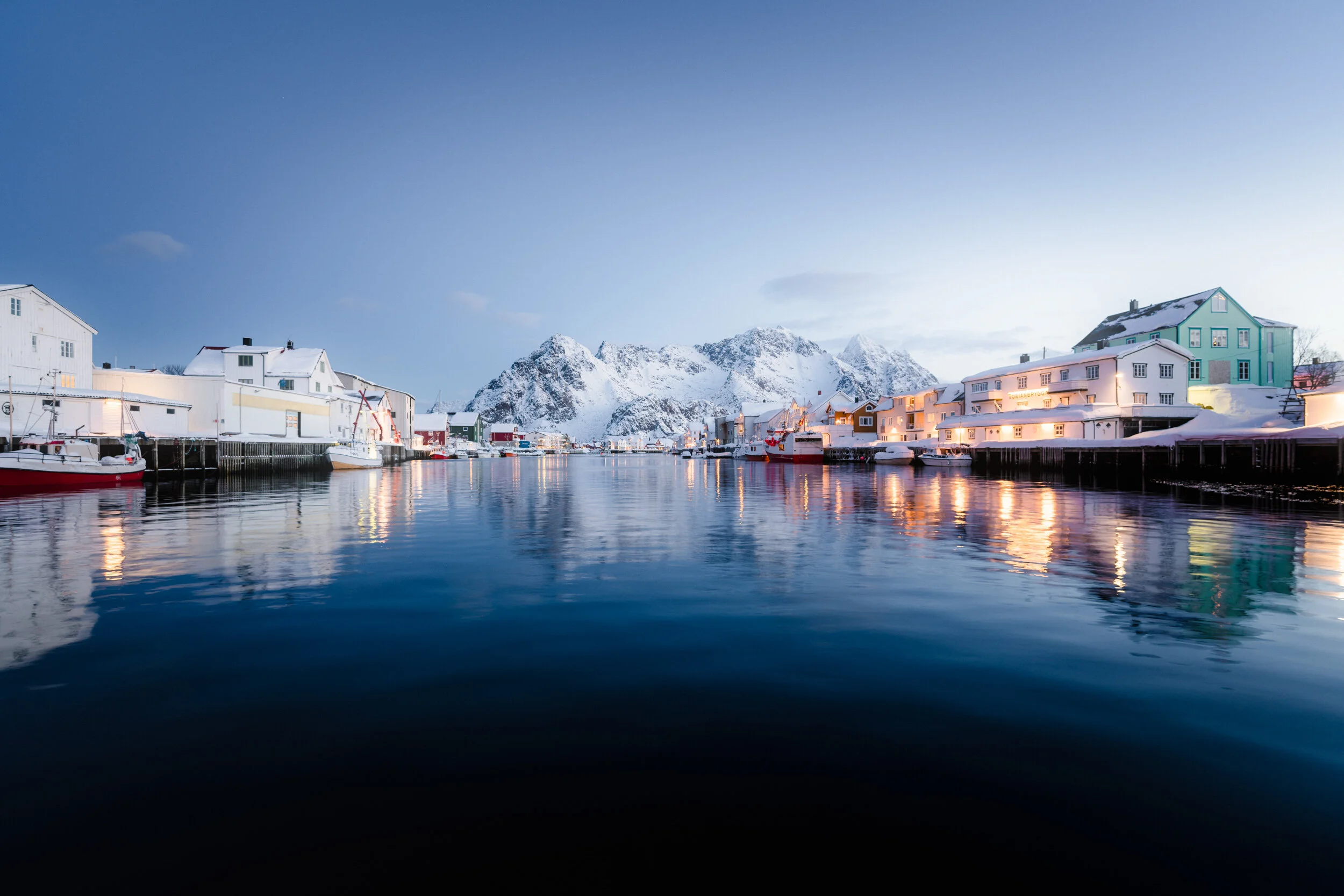Snow-covered houses along a boat-filled harbor with snowy mountains in the background during twilight.