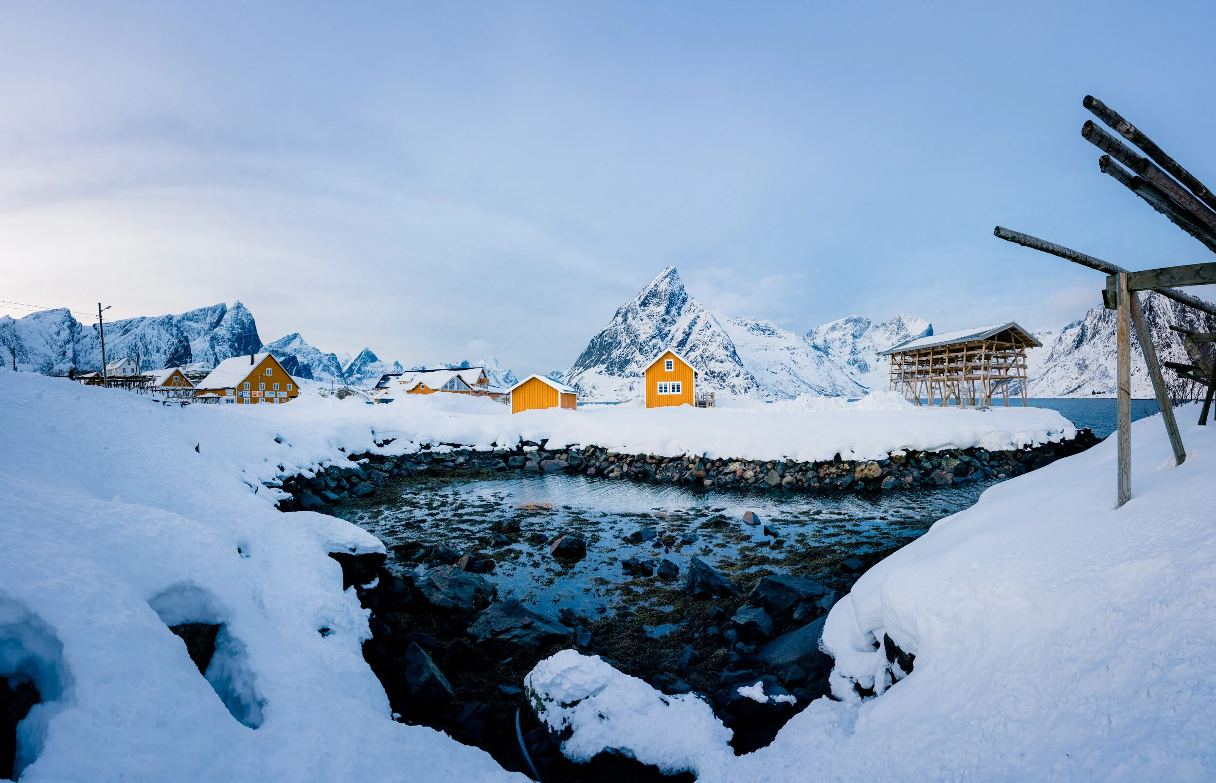 Snow-covered mountain village with yellow houses, a small river, and steep mountains in the background under a blue sky.