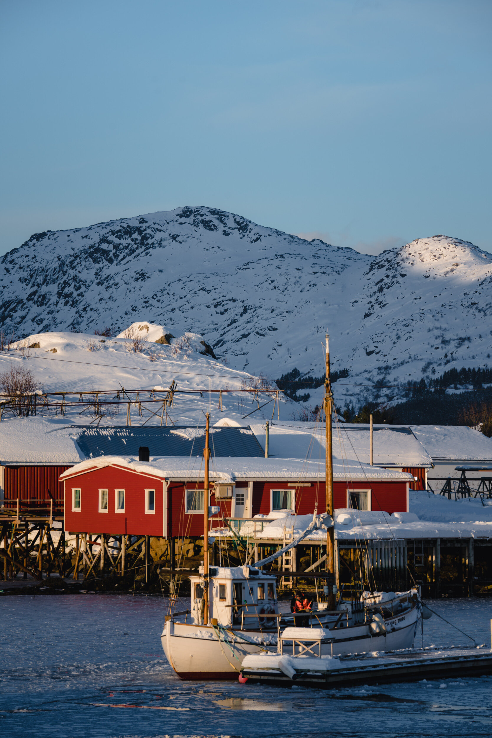 A snowy mountain landscape with a red house on stilts near the water, and a boat docked in a harbor.