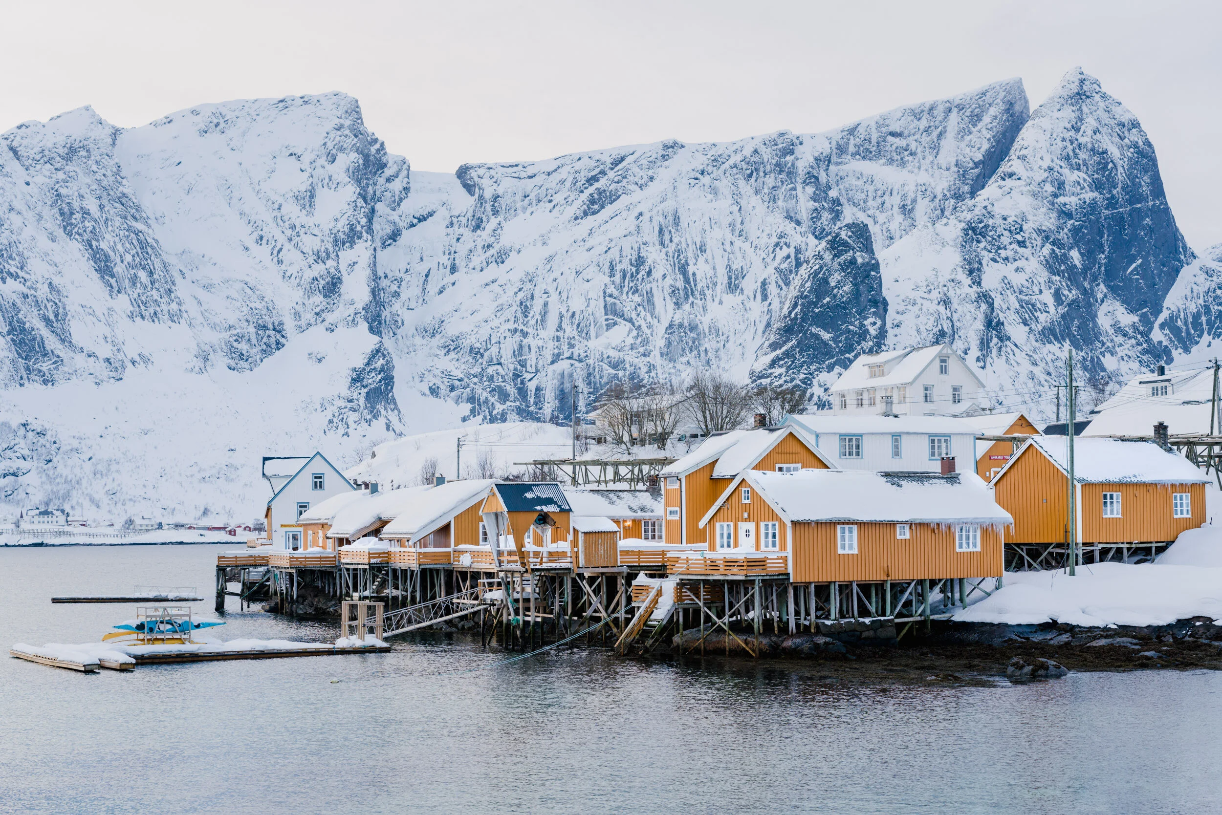 A snowy coastal village with orange houses on stilts by the water, with a background of snow-covered mountains.