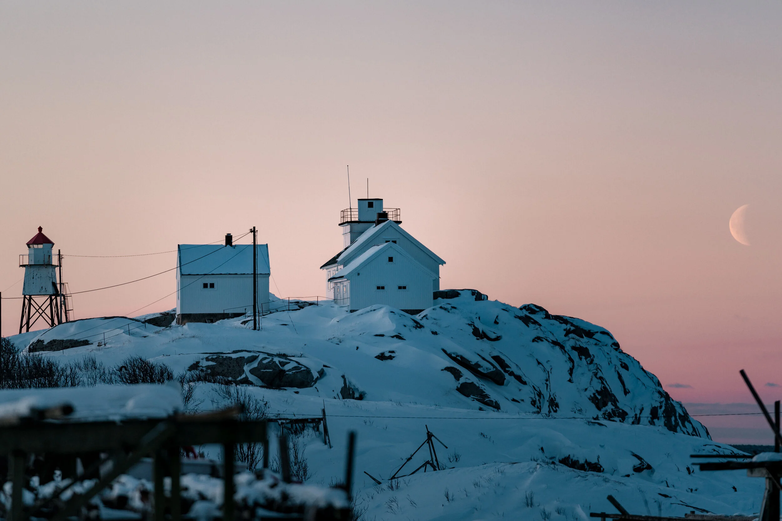 Snow-covered rocky hill with three white buildings and two lighthouses, set against a twilight sky with a visible crescent moon.