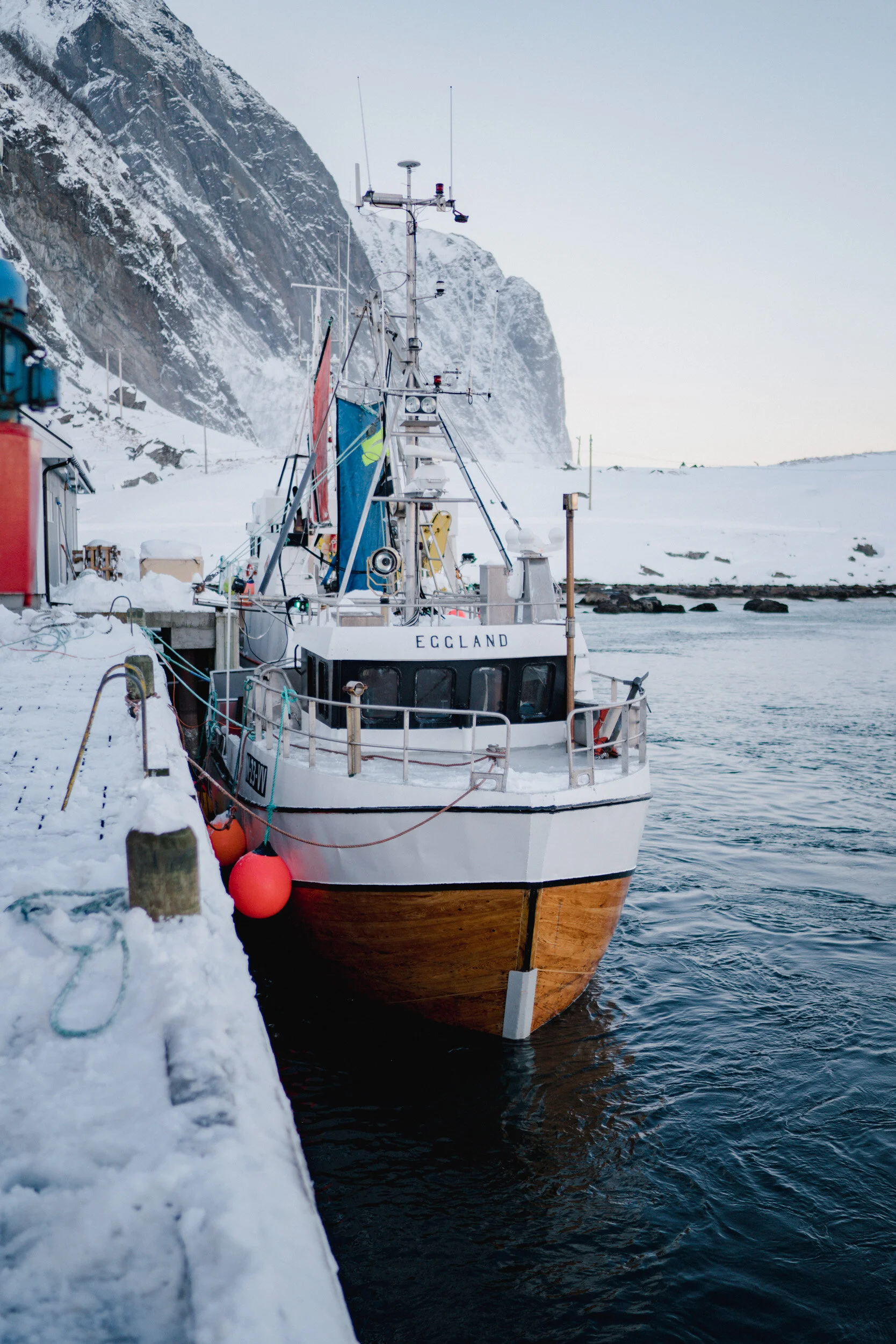 A boat docked at a snowy pier with a snow-covered mountain in the background and the water of the harbor visible.