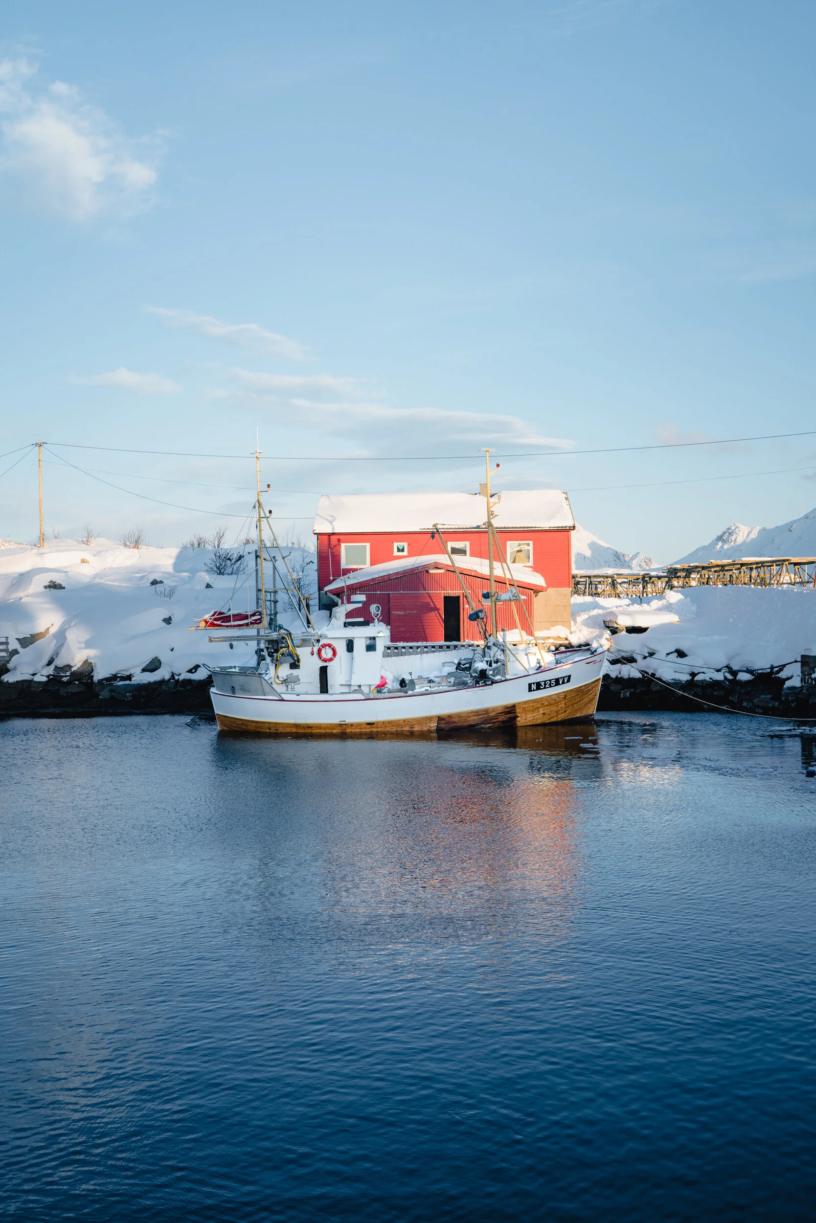 A boat floating on dark water with a red house and snow-covered landscape in the background, under a clear blue sky.