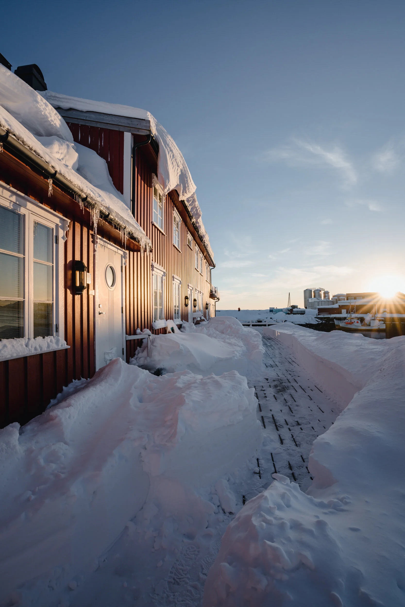 Snow-covered building exterior with icicles hanging from the roof, a snow path leading towards the horizon, and a sunset with clear sky in the background.