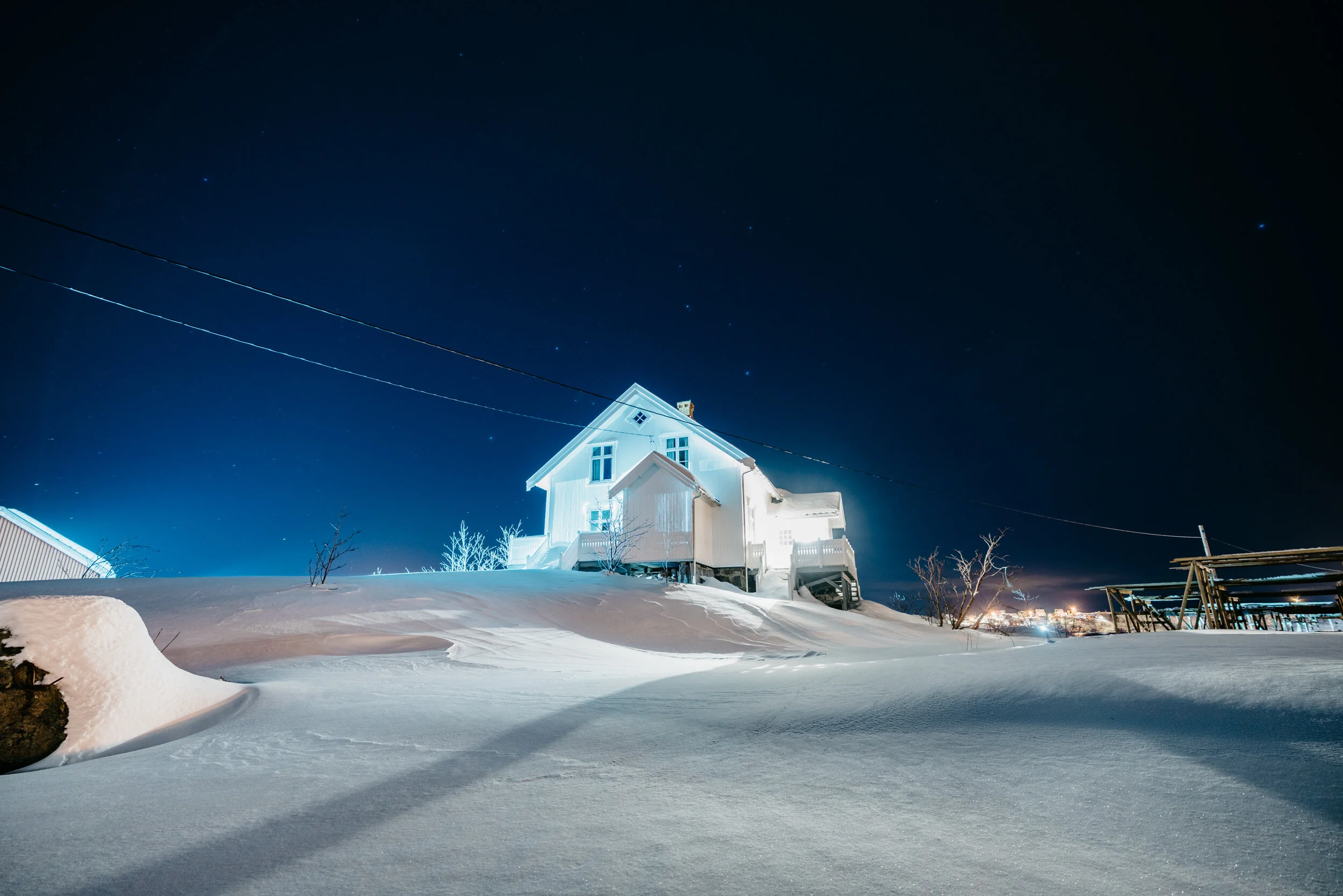 A house covered in snow at night, illuminated by exterior lights. The sky is dark with visible stars and star trails. Snow blankets the ground and surrounding area.