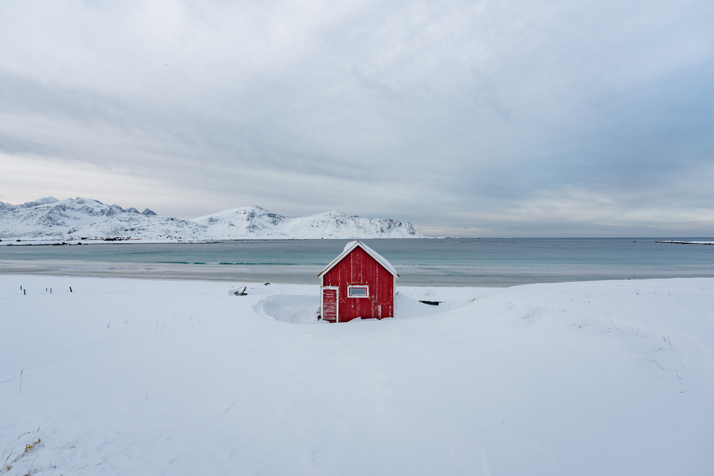 Snow-covered landscape with a small red wooden house, distant snow-capped mountains, and a body of water under a cloudy sky.