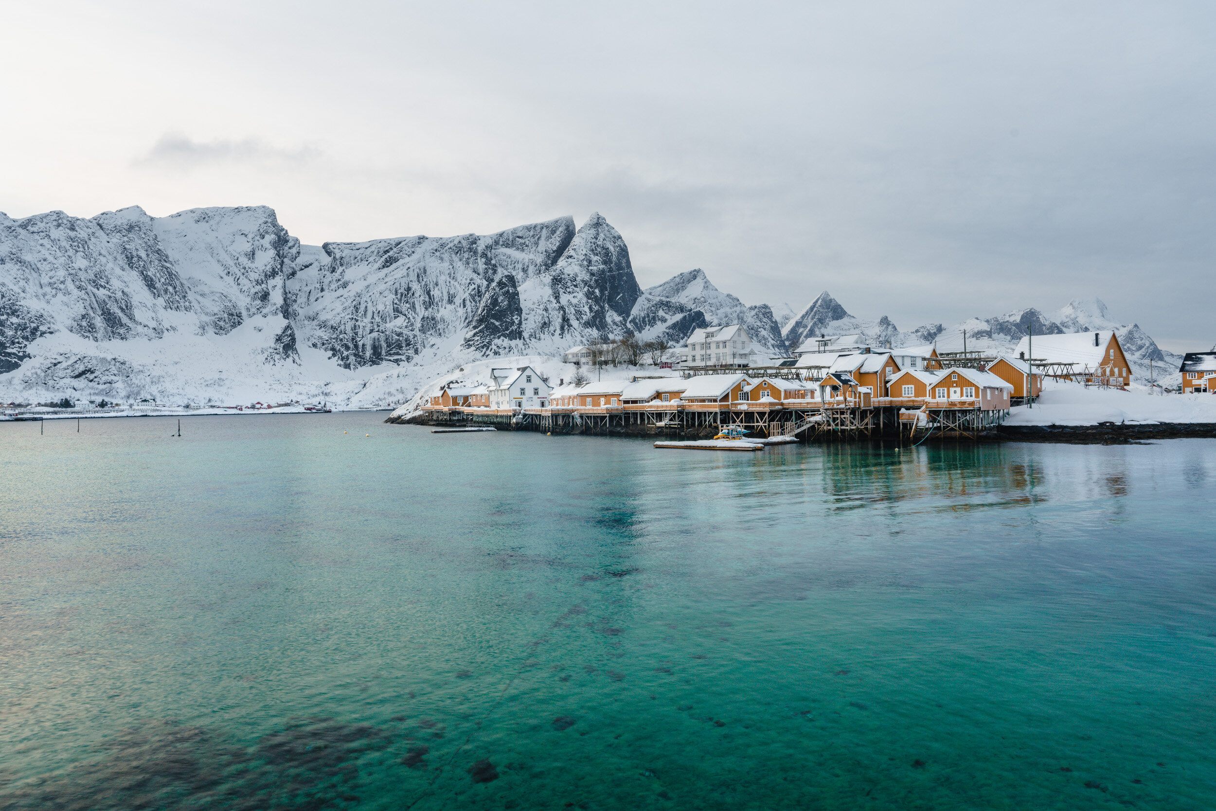 Snow-covered mountains and houses along the coastline with a calm body of water in the foreground.