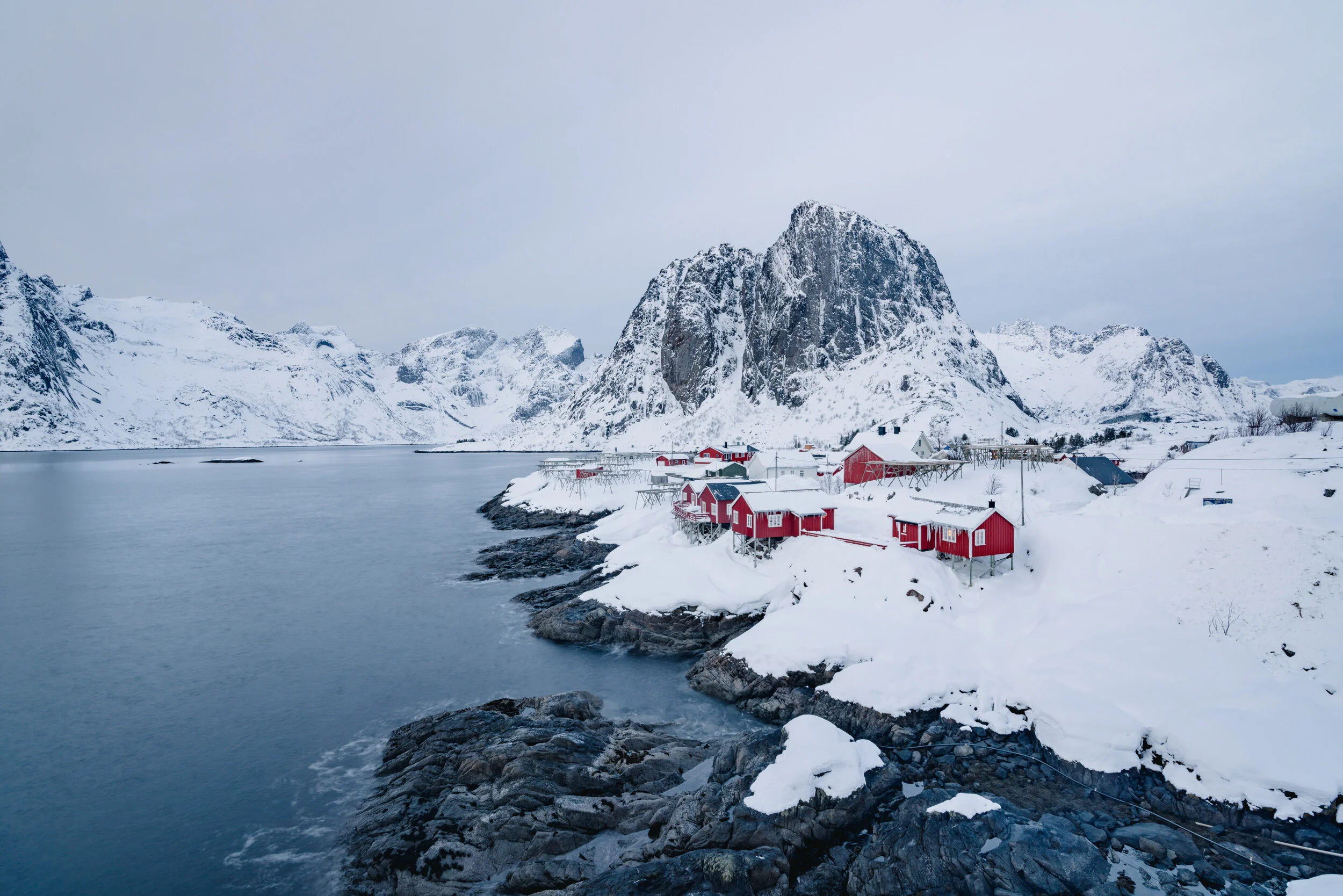 A coastal village with red houses on snow-covered ground, surrounded by icy water and snow-capped mountains under a cloudy sky.