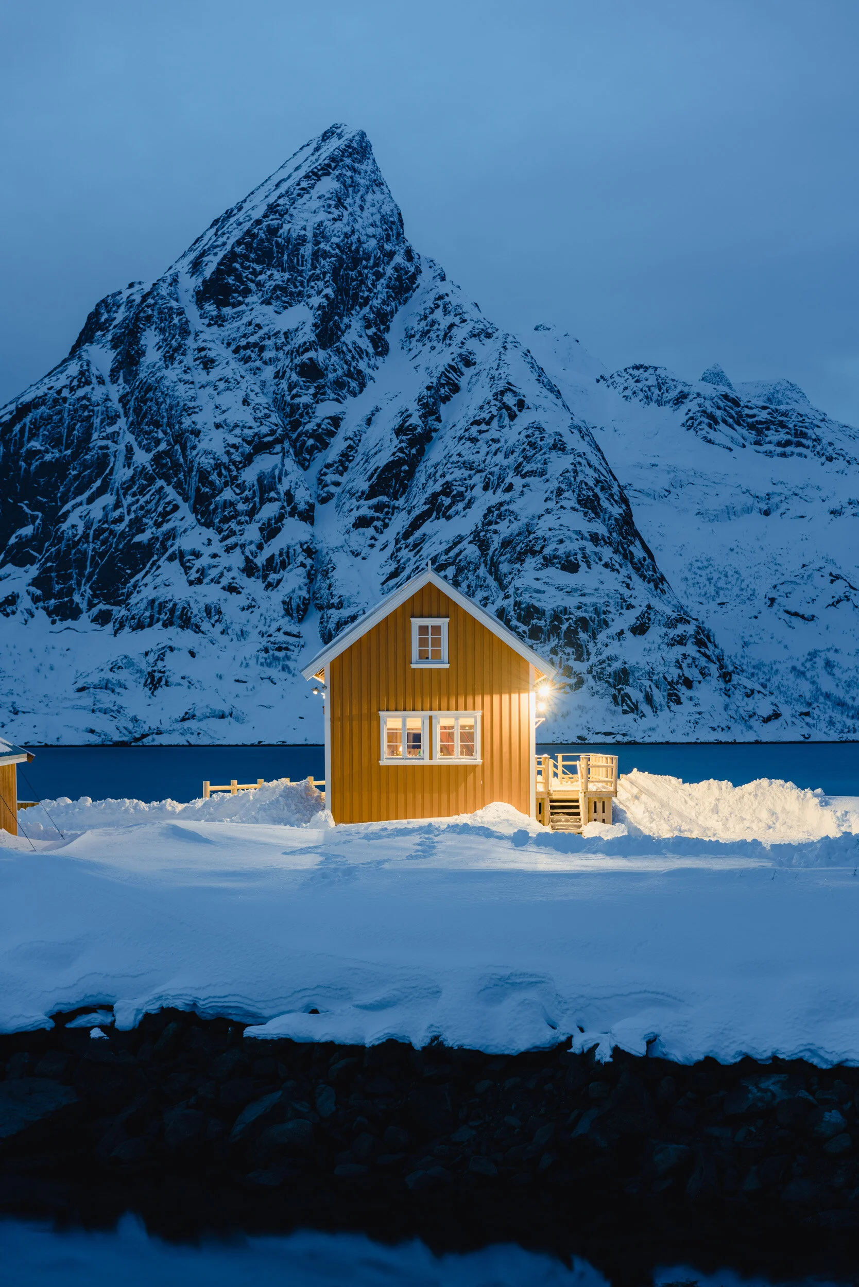 Snow-covered yellow house with lit windows in a winter landscape, mountains in the background, and a frozen body of water in the foreground.
