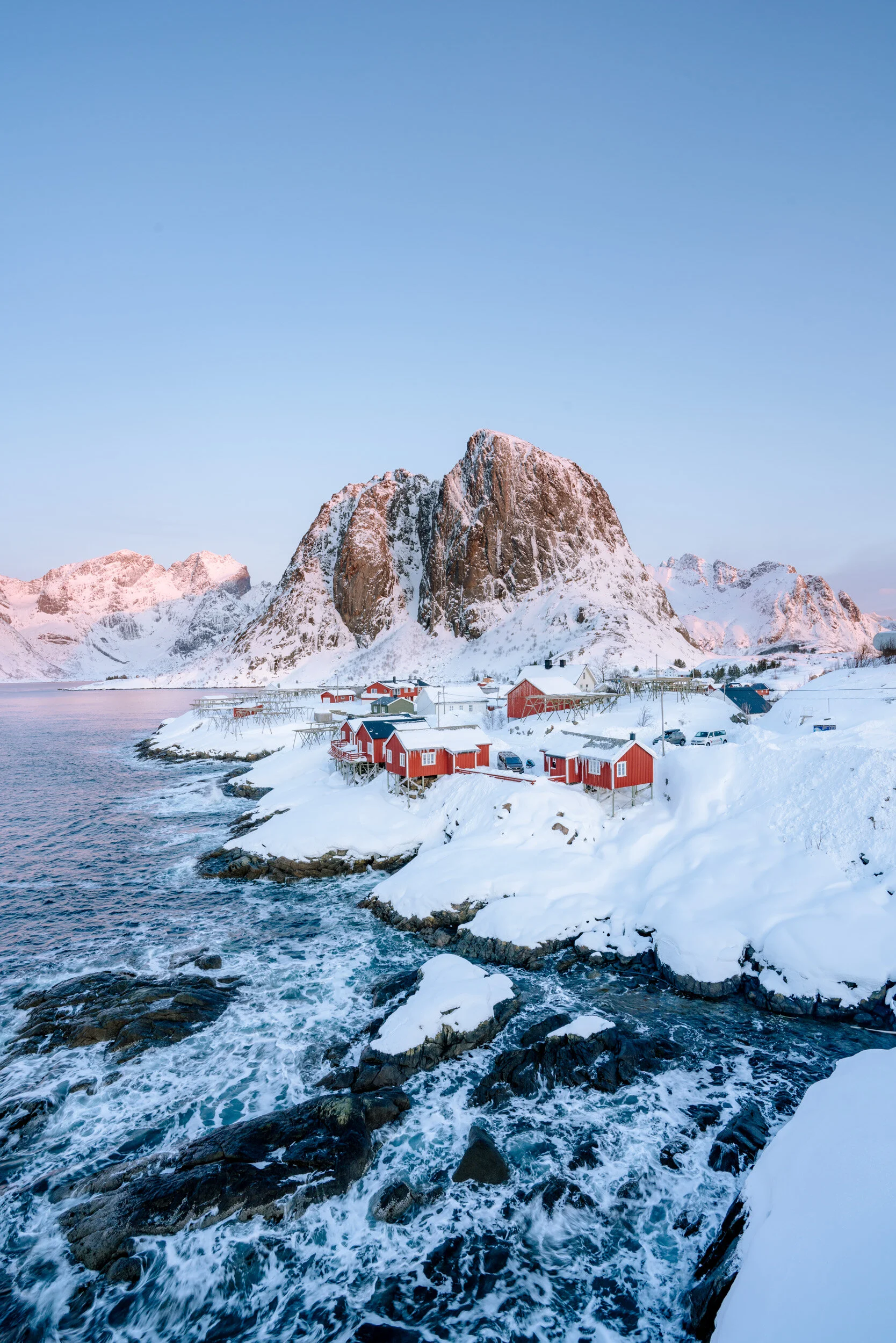 Snow-covered coastal village with red houses, rocky shoreline, and a large mountain in the background under a clear blue sky.