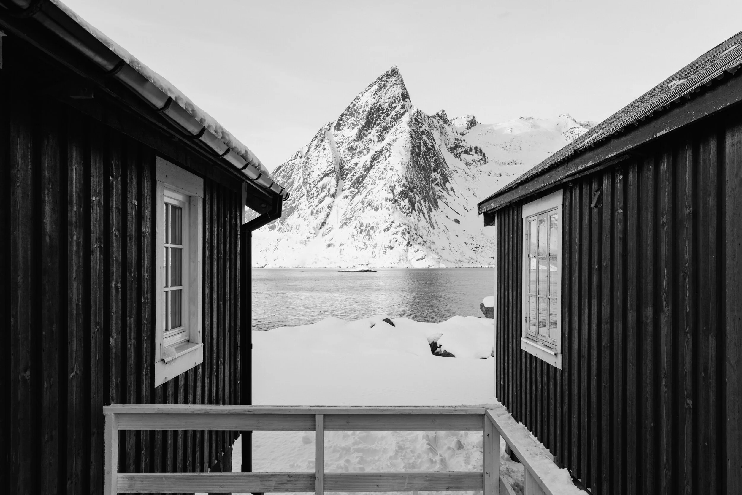 Black and white photo of a snow-covered mountain with two wooden cabins in the foreground, framed by the cabin walls on either side.