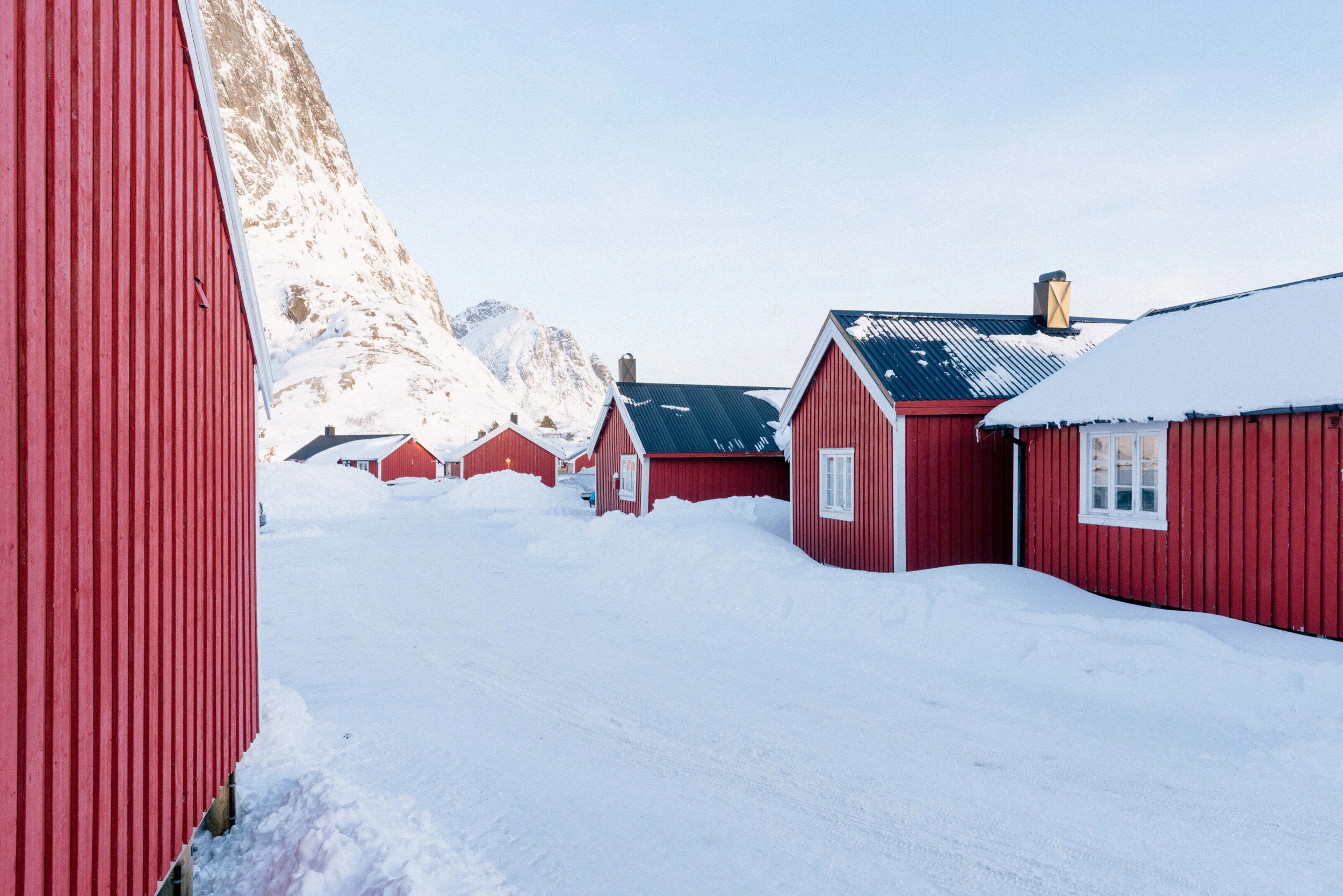 Snow-covered street lined with red wooden houses with snow on roofs and a mountainous backdrop.