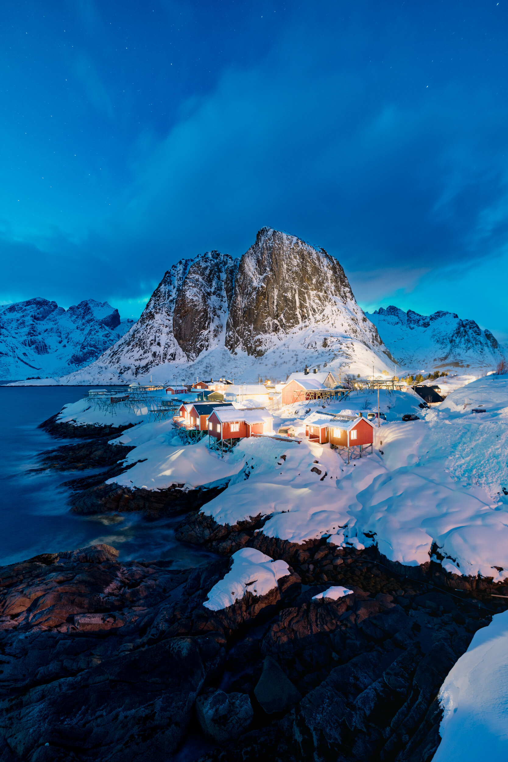 Snow-covered mountains and small red and white houses along the shoreline in a coastal village during twilight with a deep blue sky and stars.