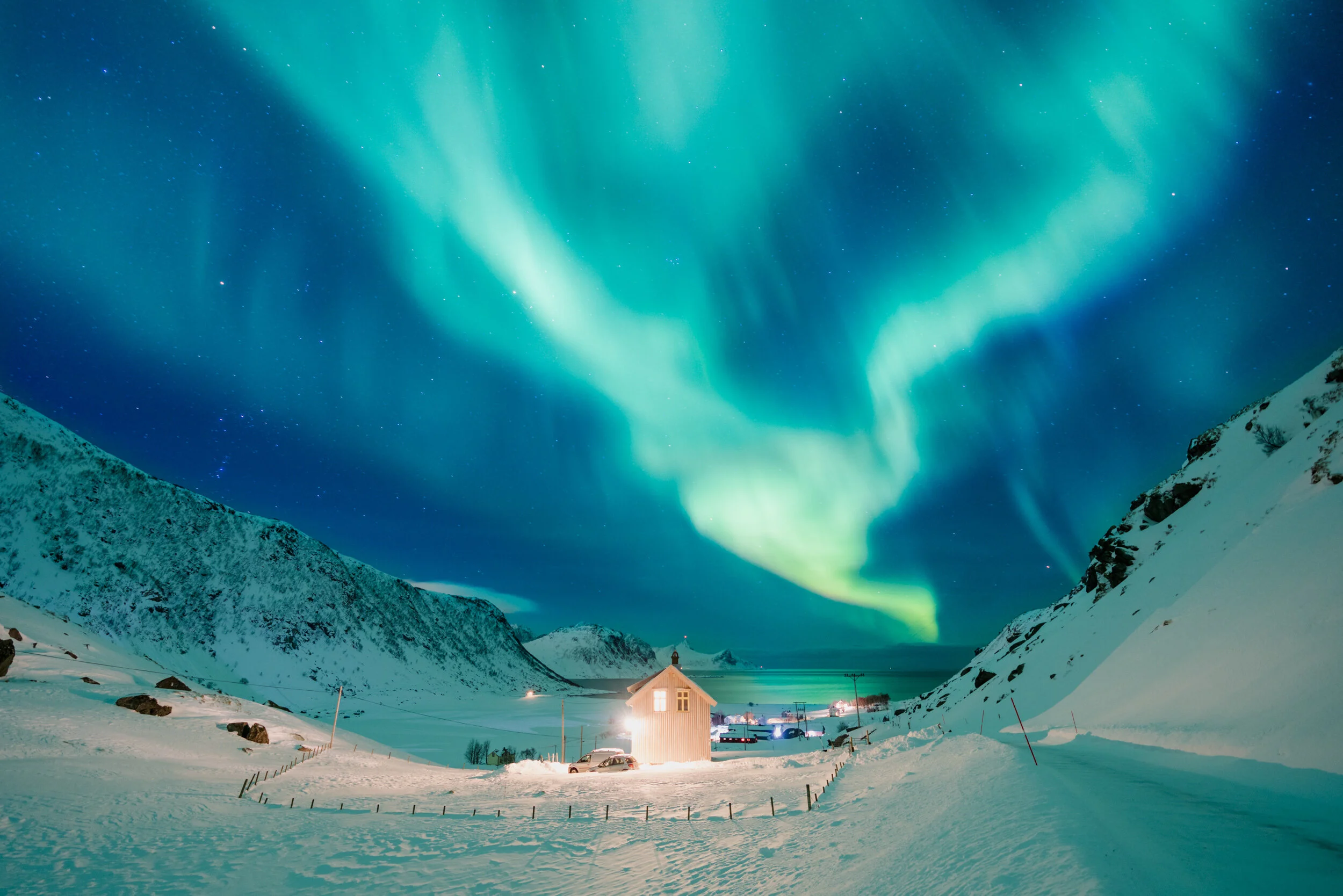 Northern lights illuminating snowy mountains and a small house in a winter landscape at night.