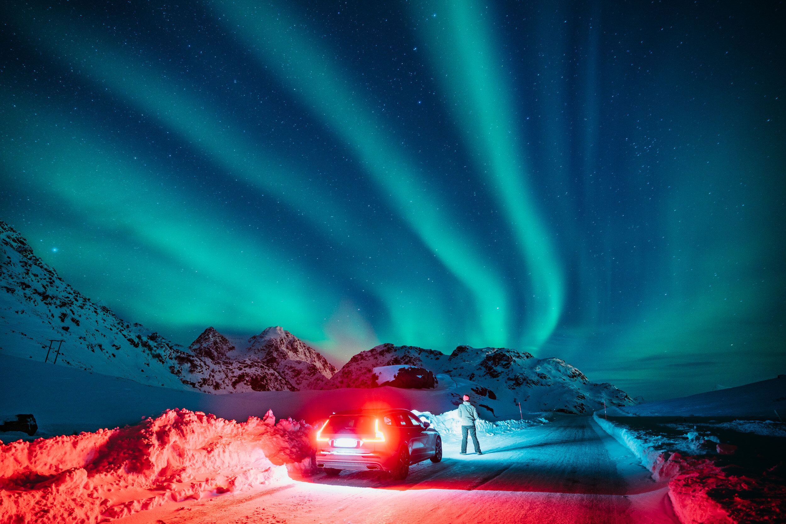 Night sky illuminated by Northern Lights over snow-covered mountains and a snow-packed road. A person stands beside a car with bright taillights shining.