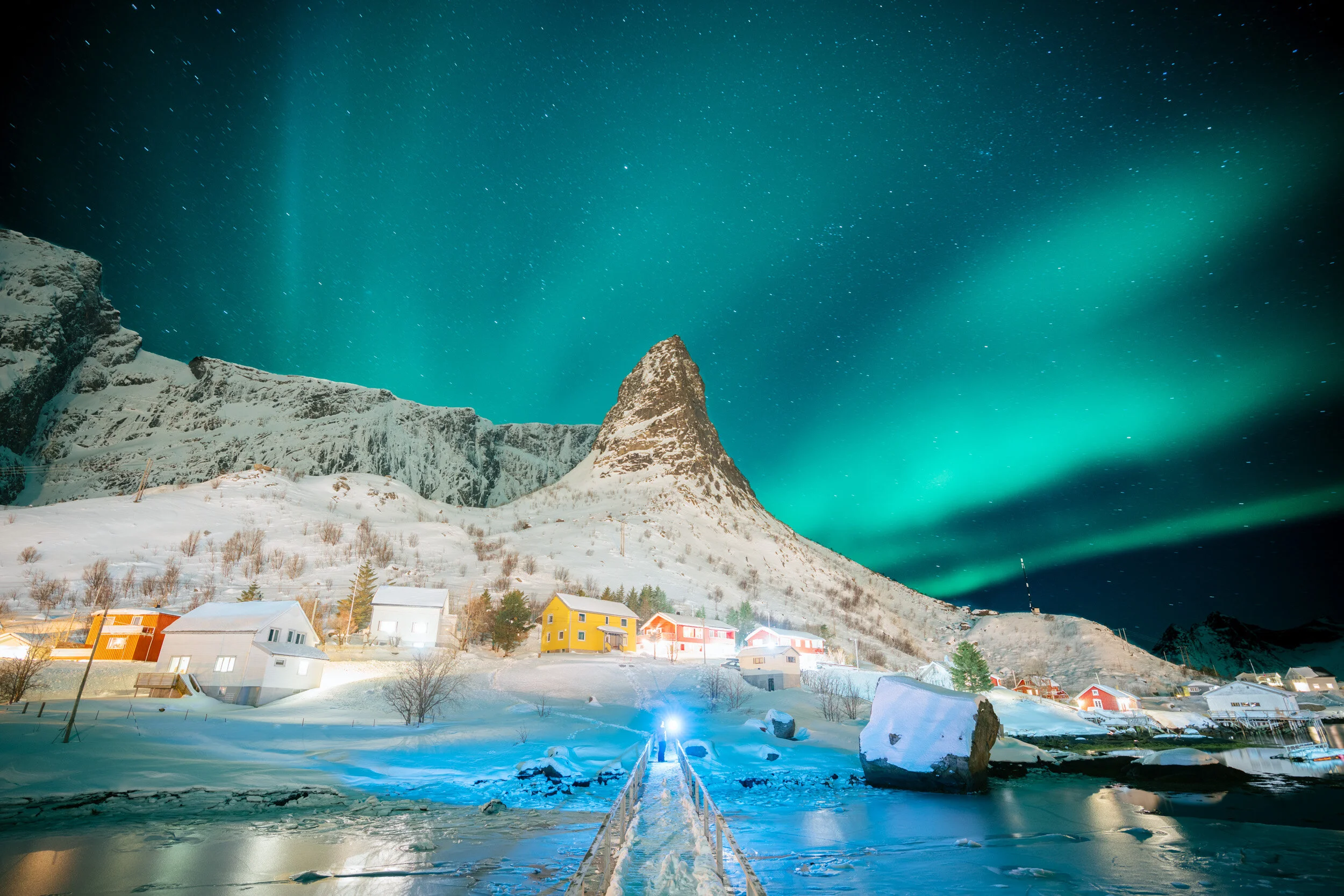 Nighttime view of a snowy Norwegian village with colorful houses, mountains, Northern Lights, and a starry sky.
