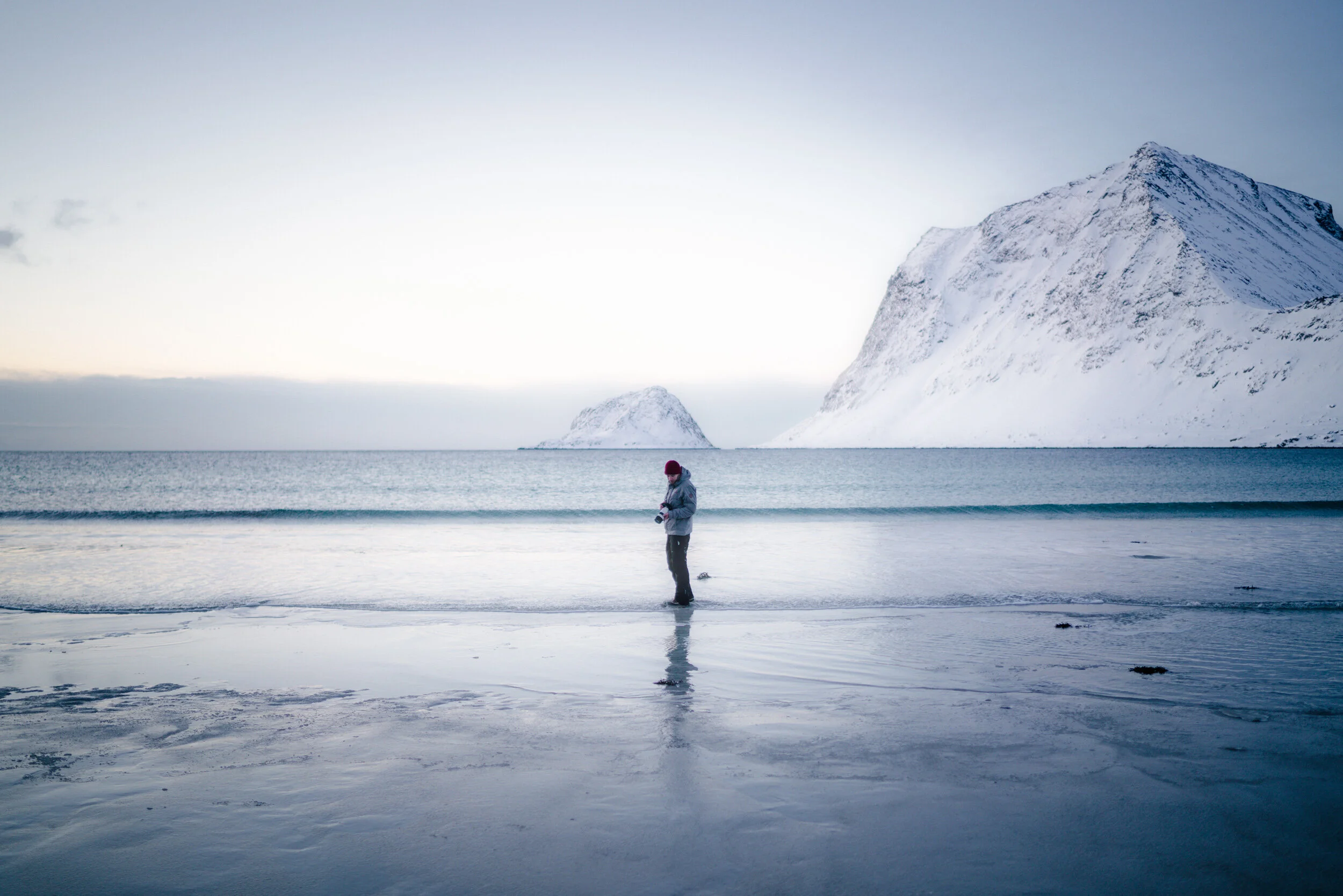 A person standing on a sandy, snowy beach near the water, with snow-covered mountains in the background under a cloudy sky.