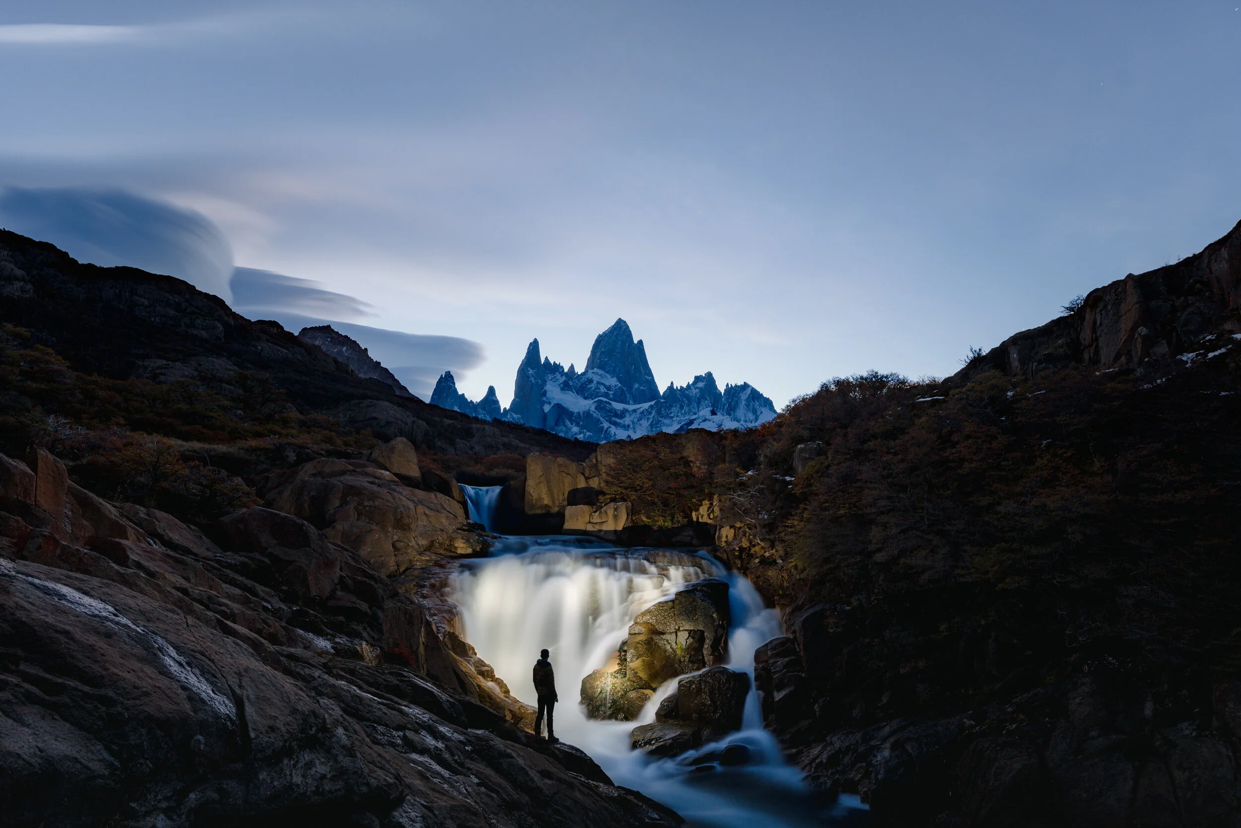 Person standing on rocks near a waterfall with mountains in the background under a cloudy sky.