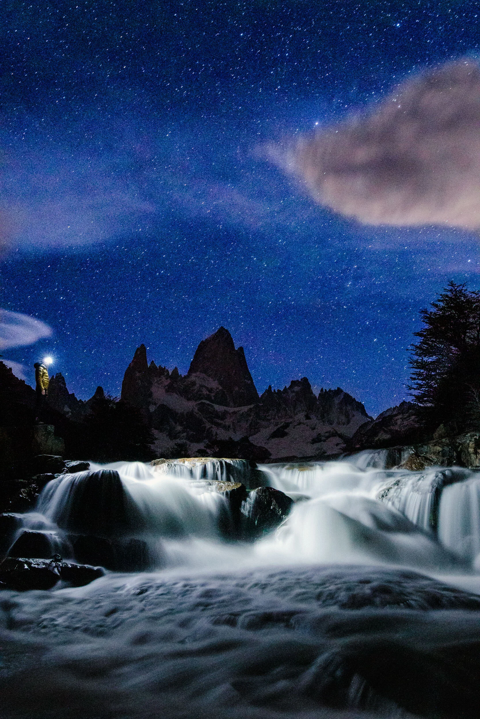Night sky full of stars above rugged mountain peaks and flowing river in a remote wilderness.