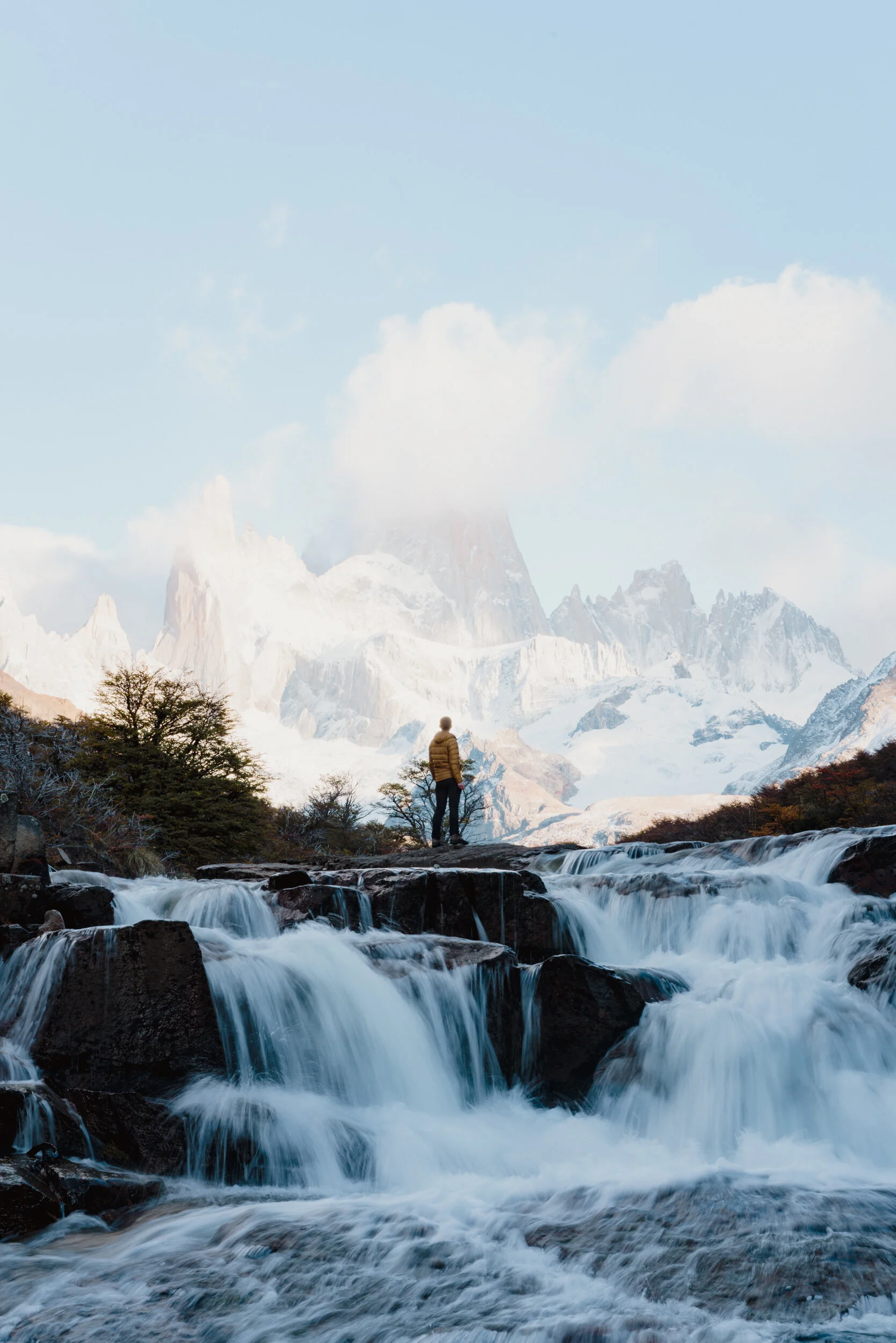 A person standing on rocks near cascading river waters with snow-capped mountains in the background under a partly cloudy sky.