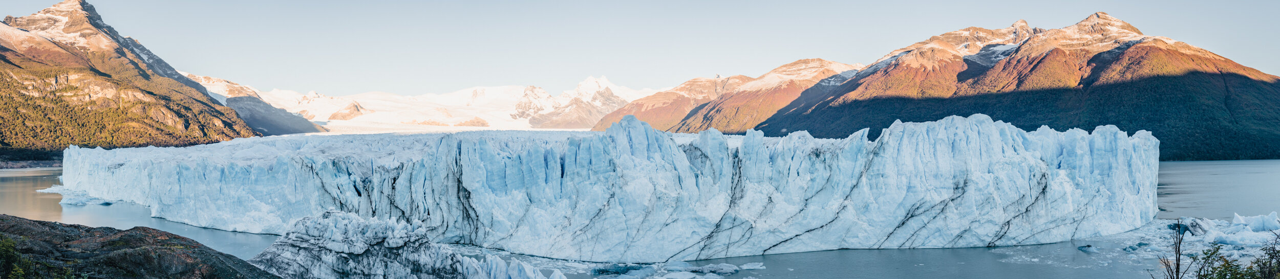 A large glacier with jagged ice facing a calm body of water, surrounded by mountains with some snow-capped peaks under a clear sky.