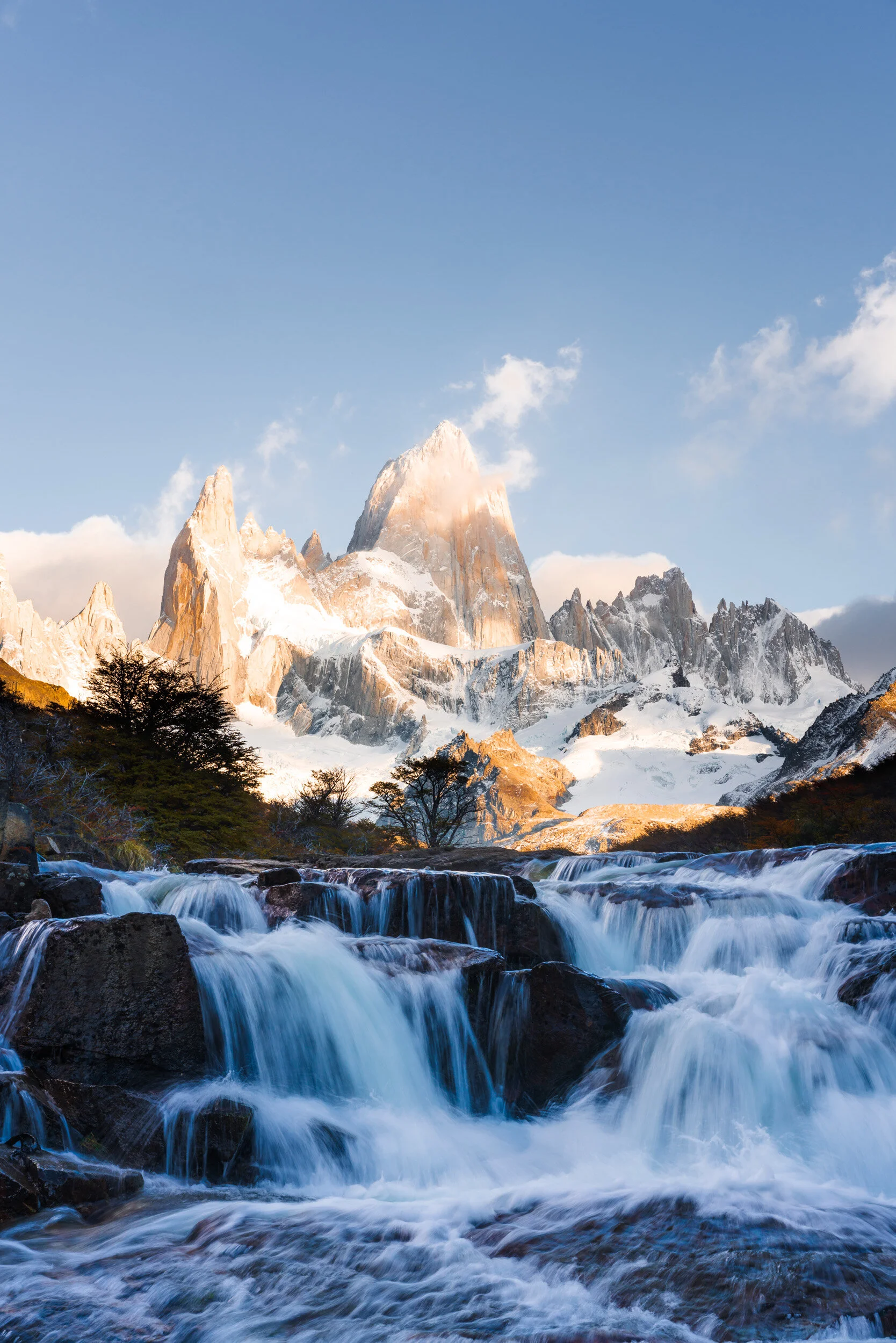 A mountain landscape with snow-capped peaks, a flowing river in the foreground, and a blue sky with some clouds.
