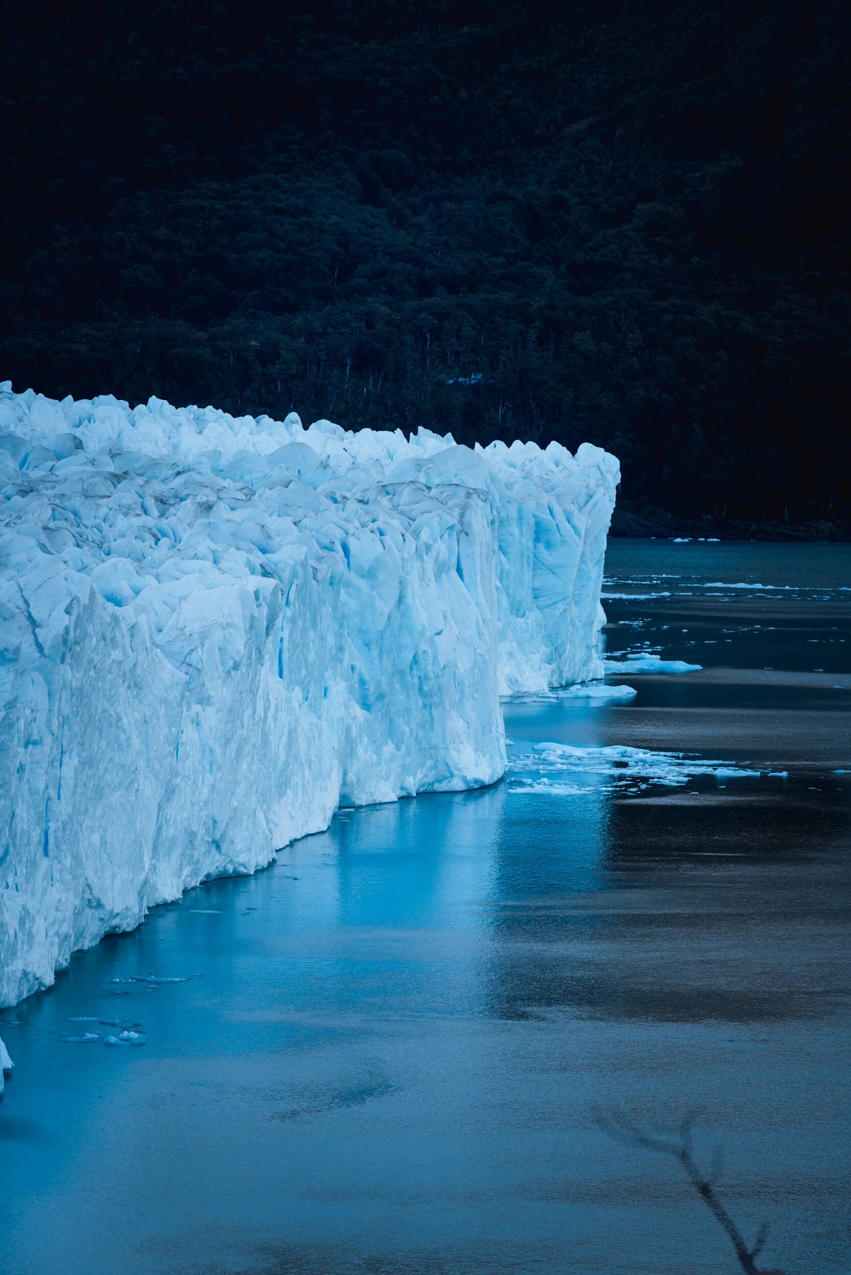 A glacier with jagged white ice formations over dark water, surrounded by a forested mountain landscape at dusk.