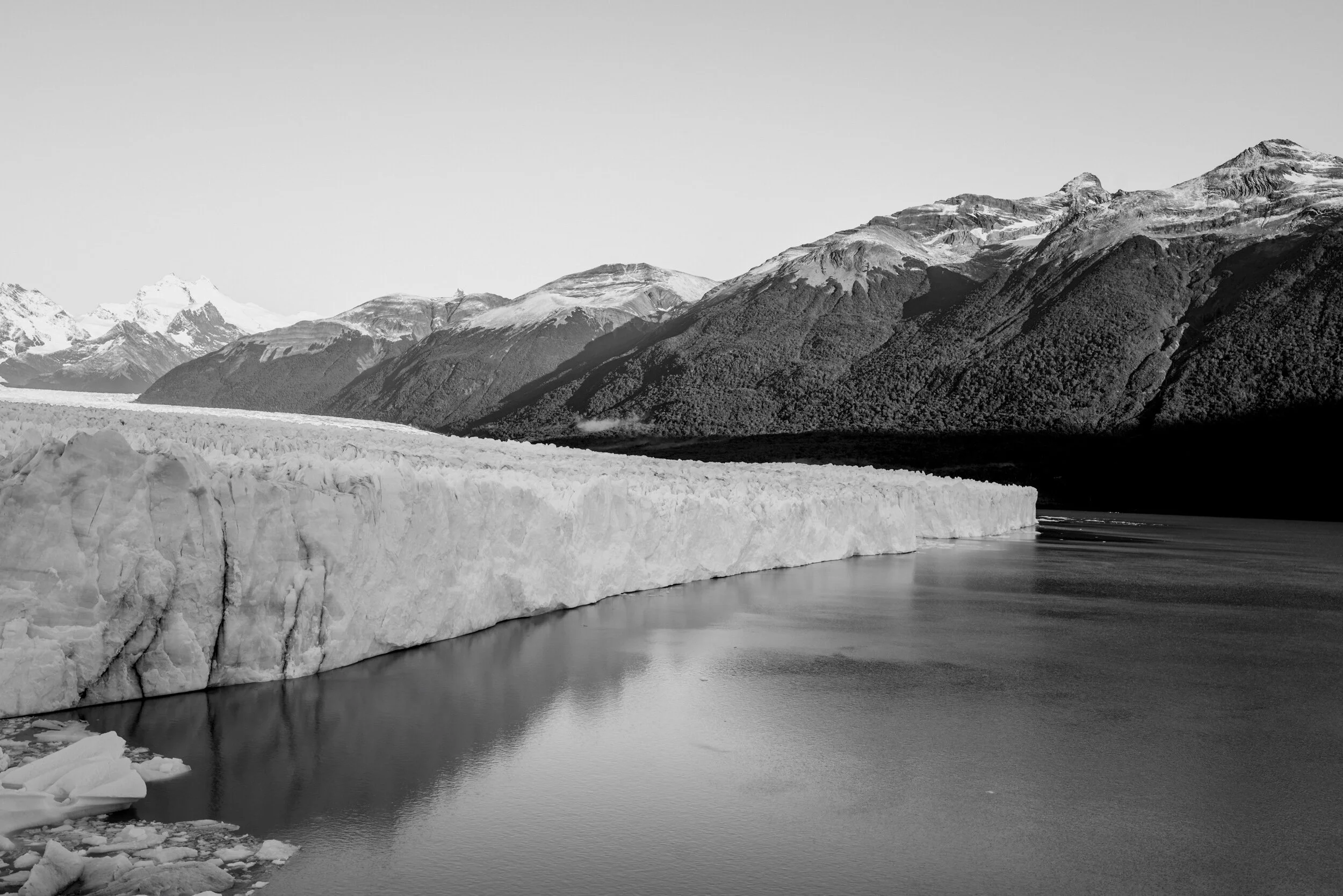 Black and white photo of a glacier with snow-capped mountains in the background and calm water in the foreground.