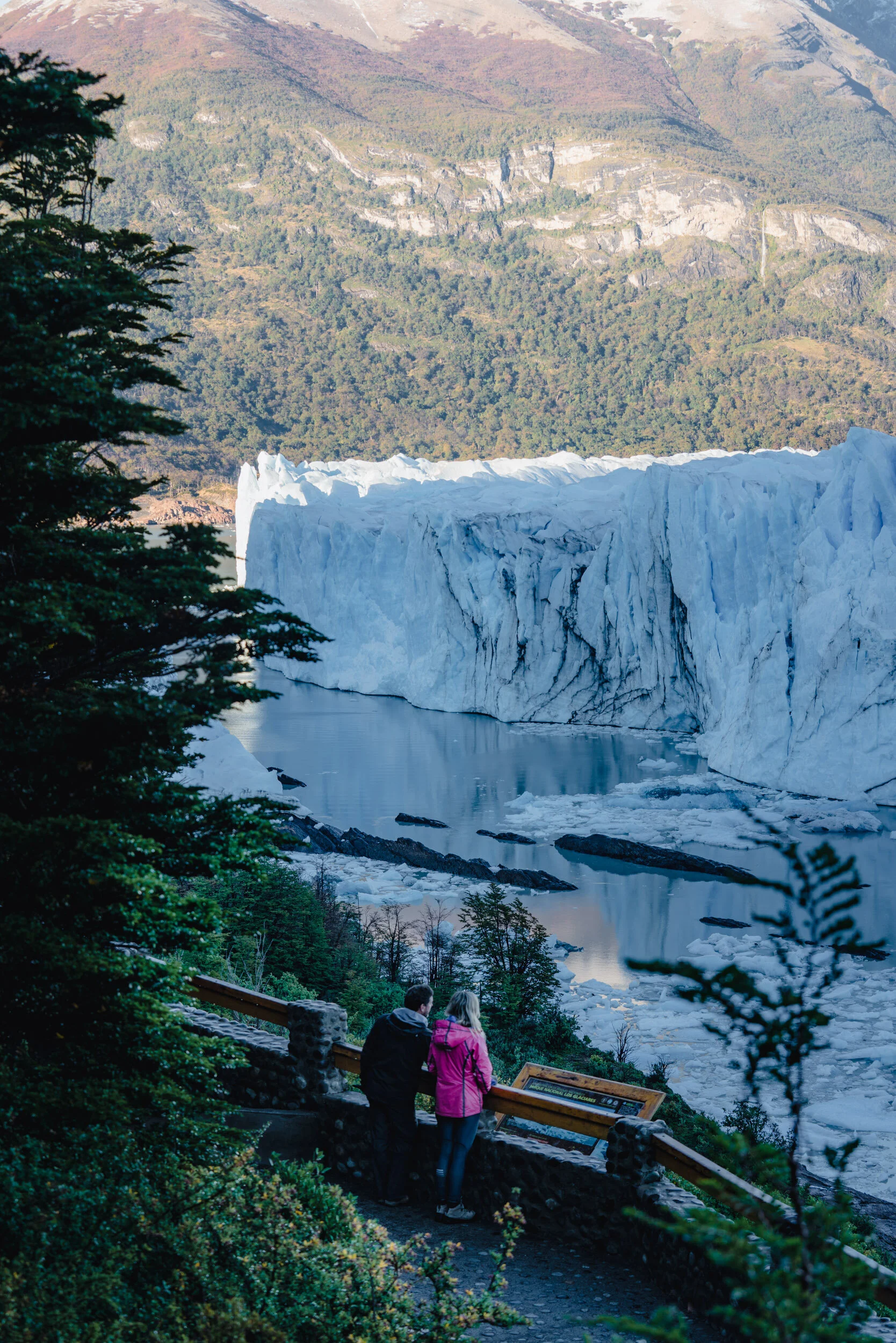 Two people, a man and a woman, stand near a scenic overlook of a glacier and mountain landscape. They are dressed in outdoor clothing and are looking at the view.