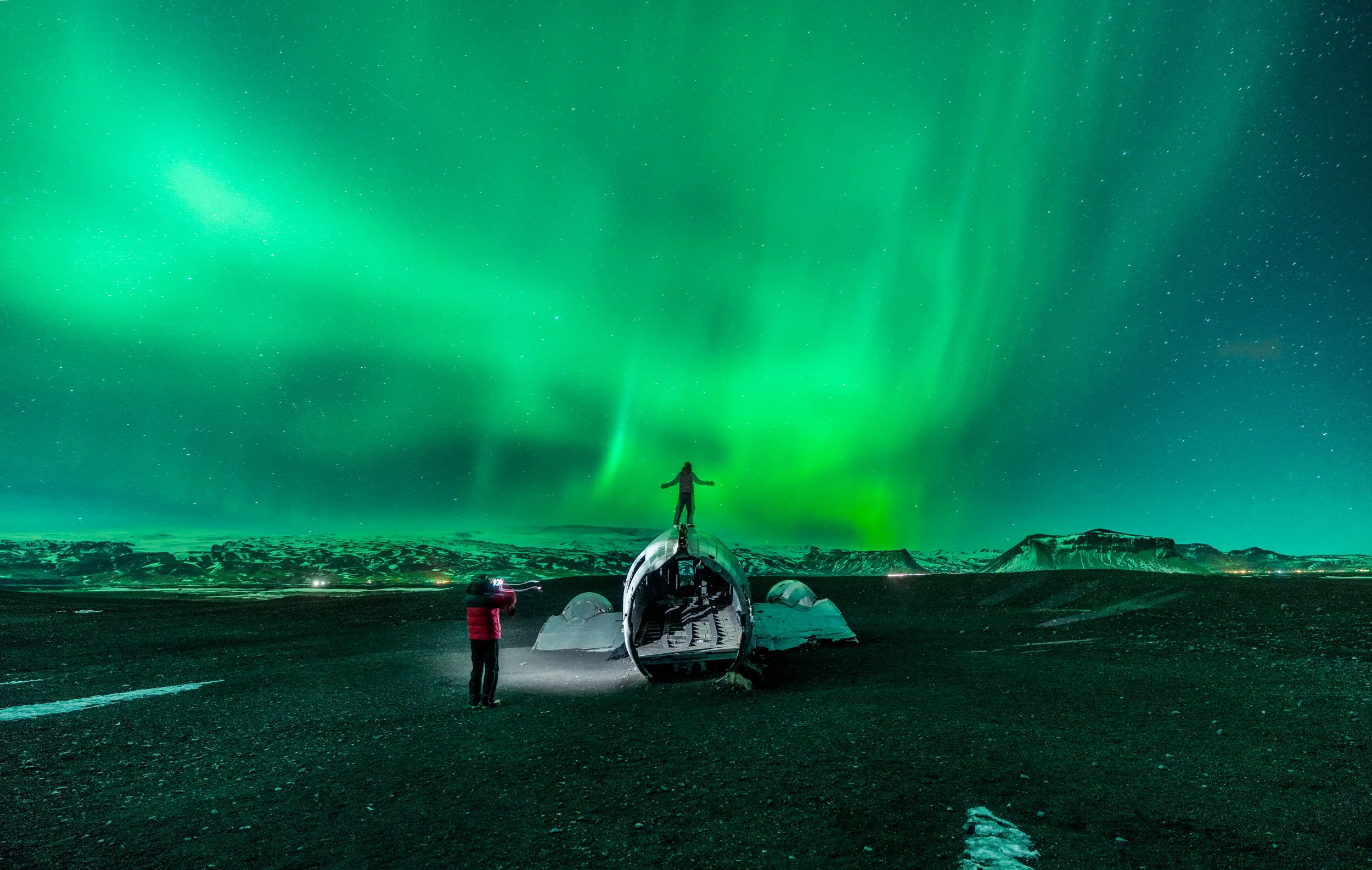 Person standing on a snowy landscape at night, watching the Northern Lights in the sky, with a small aircraft on the ground and another person nearby, capturing the scene.