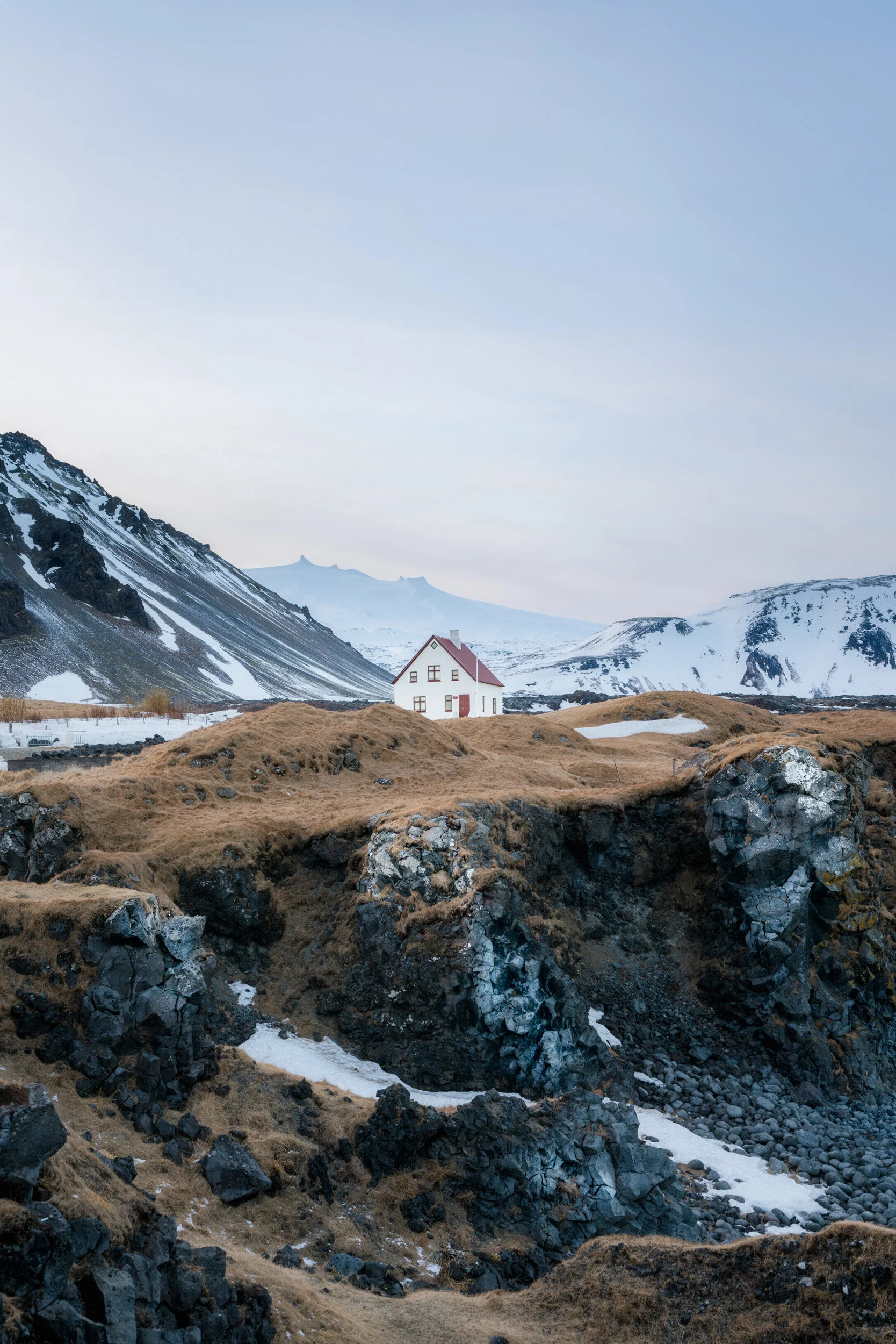 A snowy mountain landscape with a isolated white house with a red roof in a valley surrounded by dark rocky slopes and partially snow-covered ground.