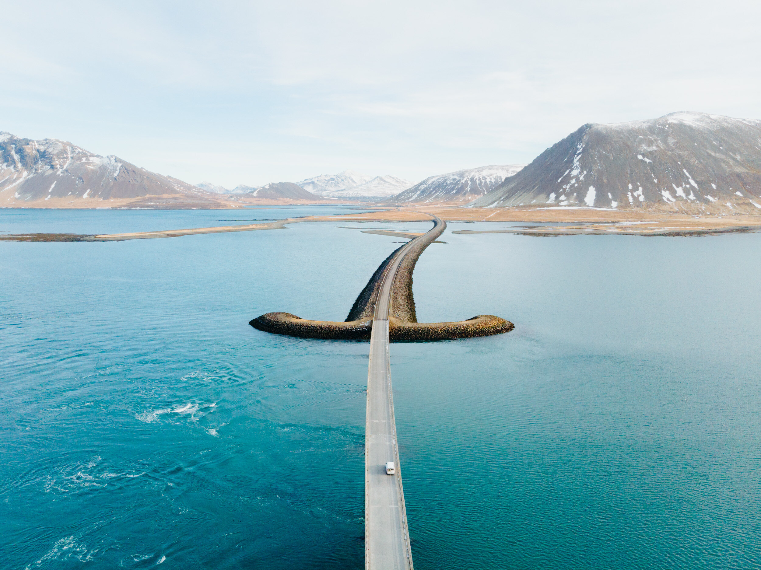 A bridge extends over a body of water, connecting to a narrow strip of land with mountains in the background, under a partly cloudy sky.