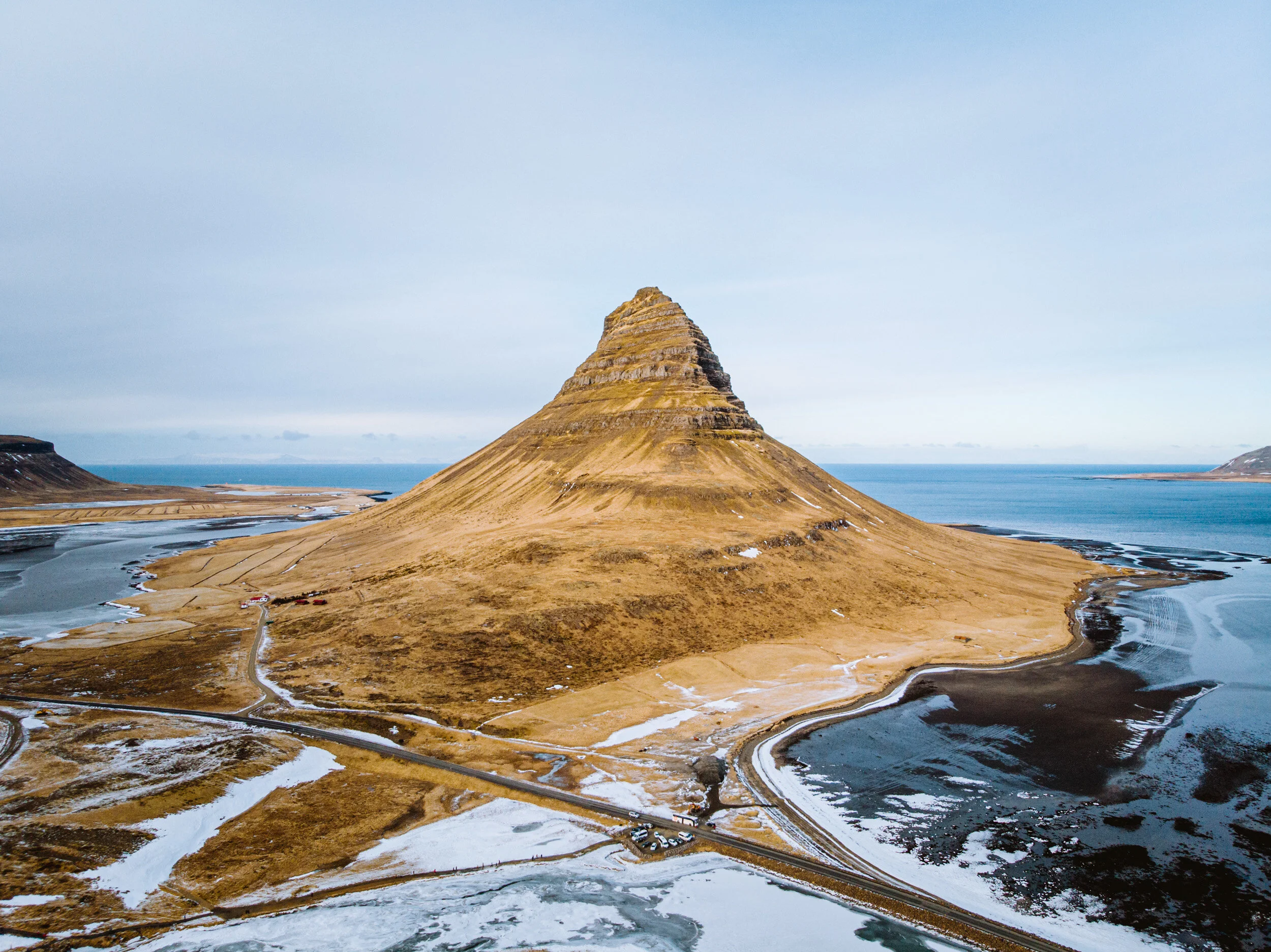 A large, conical mountain with tiered layers, surrounded by a landscape of snow patches, roads, and sparse vegetation, with a body of water in the background under a partly cloudy sky.