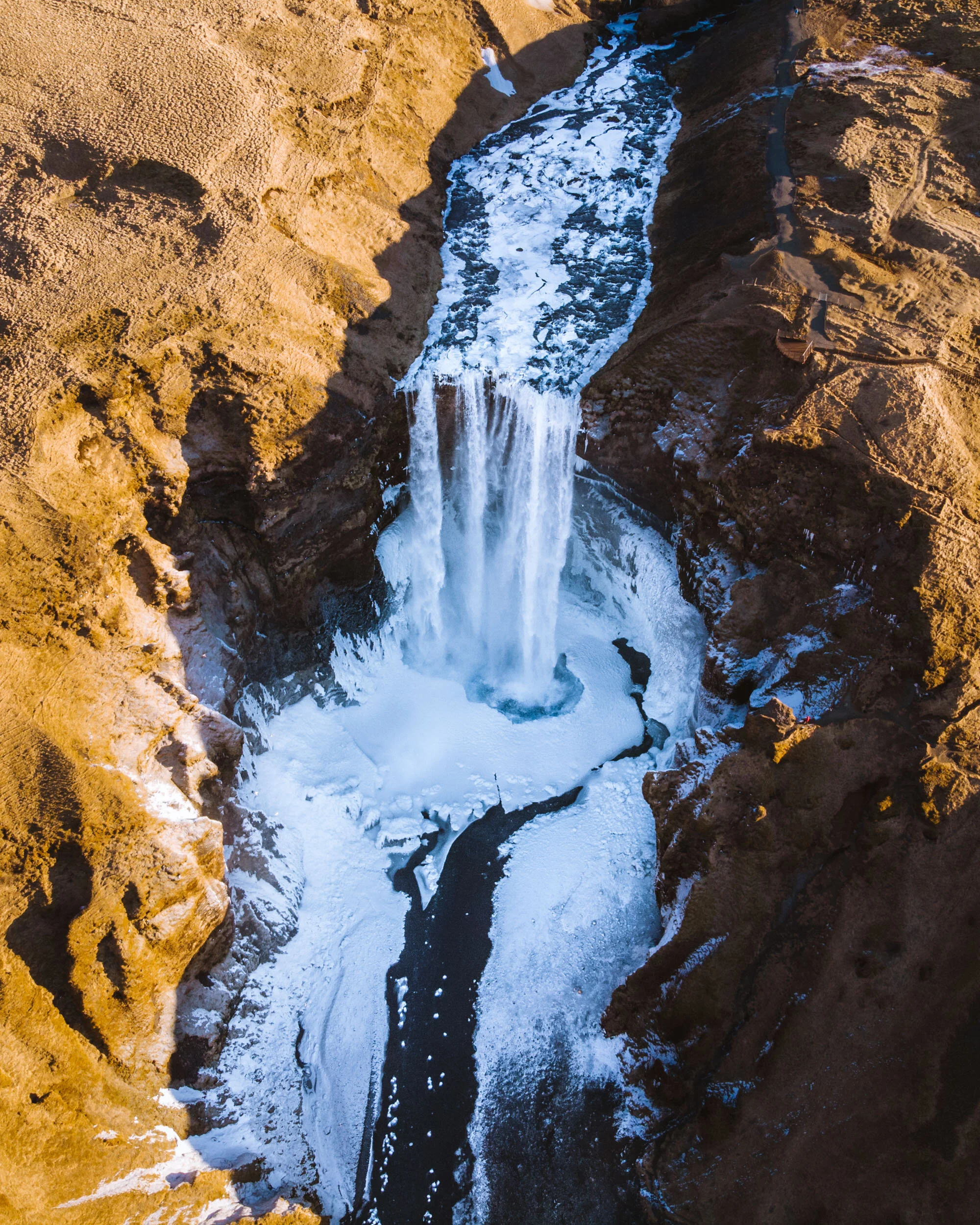 A waterfall flowing into a river in a canyon, with snow on the ground and surrounding rocky cliffs.