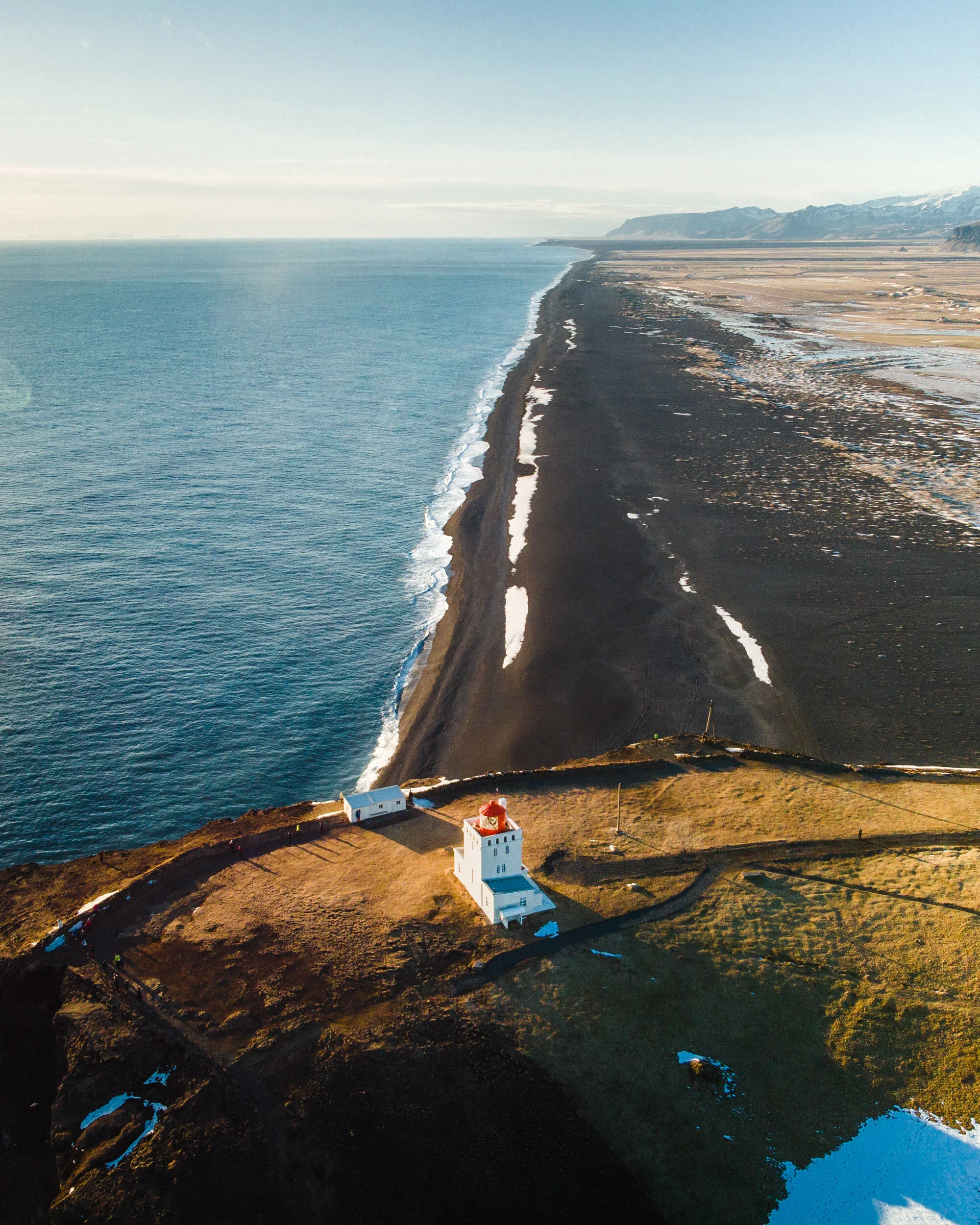 Aerial view of a black sand beach with a white lighthouse on a grassy hill, ocean to the left, mountains in the background, and patches of snow on the ground.