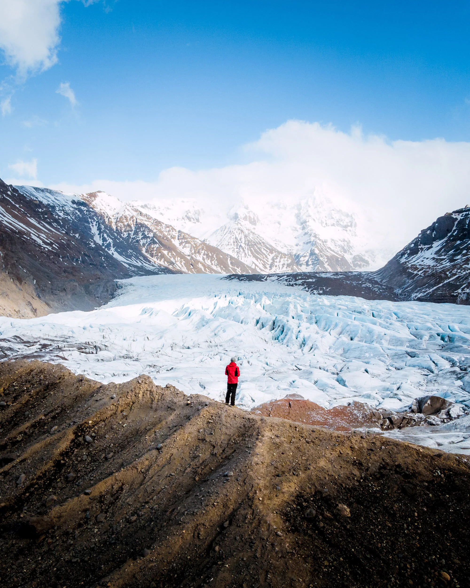 A person in a red jacket and black pants standing on rocky ground, looking at snow-covered mountains and glaciers under a partly cloudy blue sky.