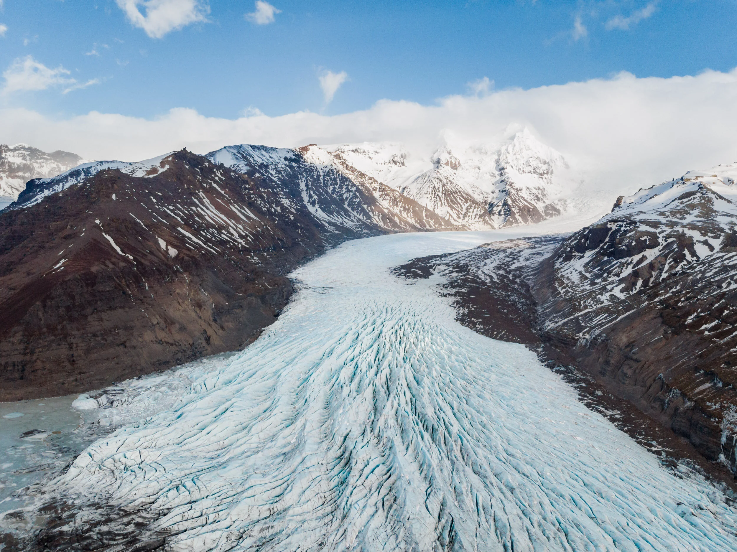 Snow-capped mountains with a glacier flowing through a valley under a partly cloudy sky.