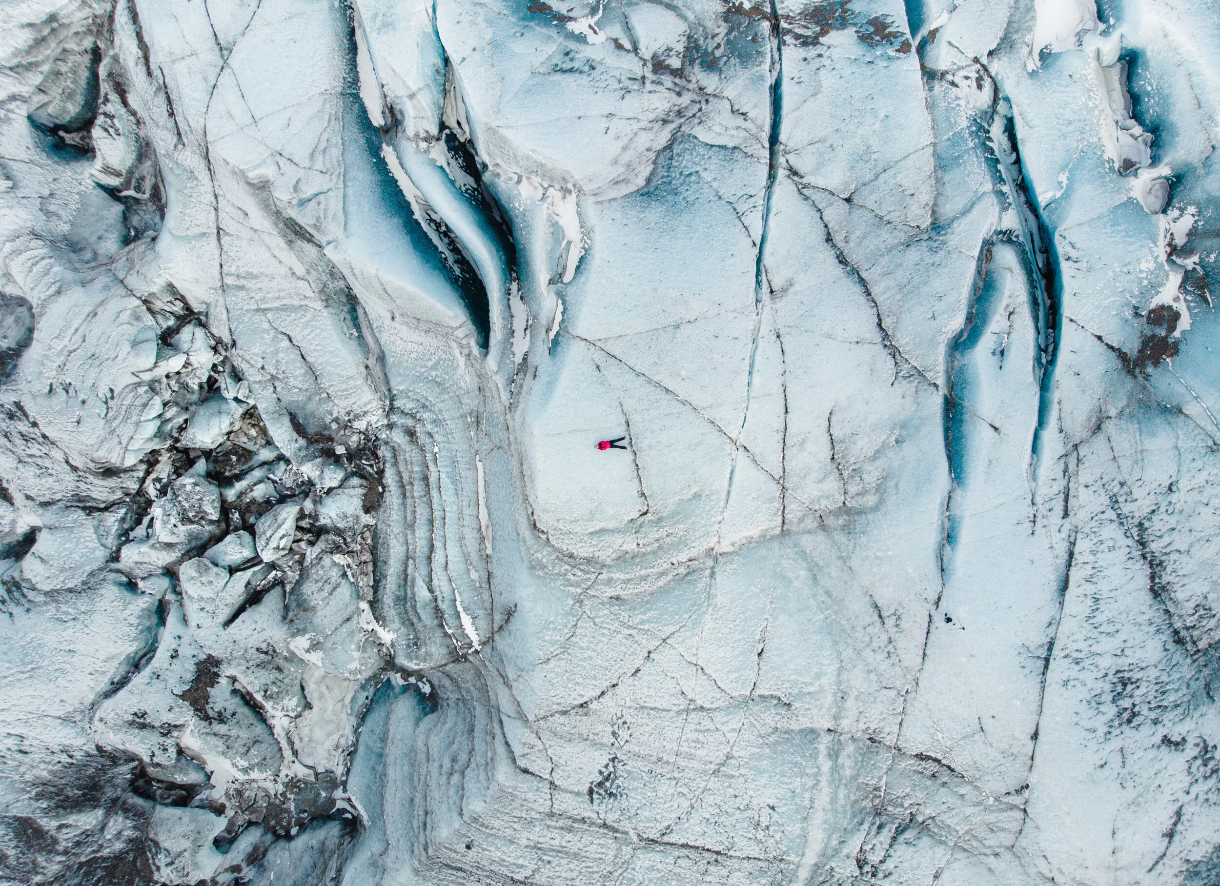 An aerial view of a glacier with deep crevasses and a person in a bright pink jacket standing on the ice.