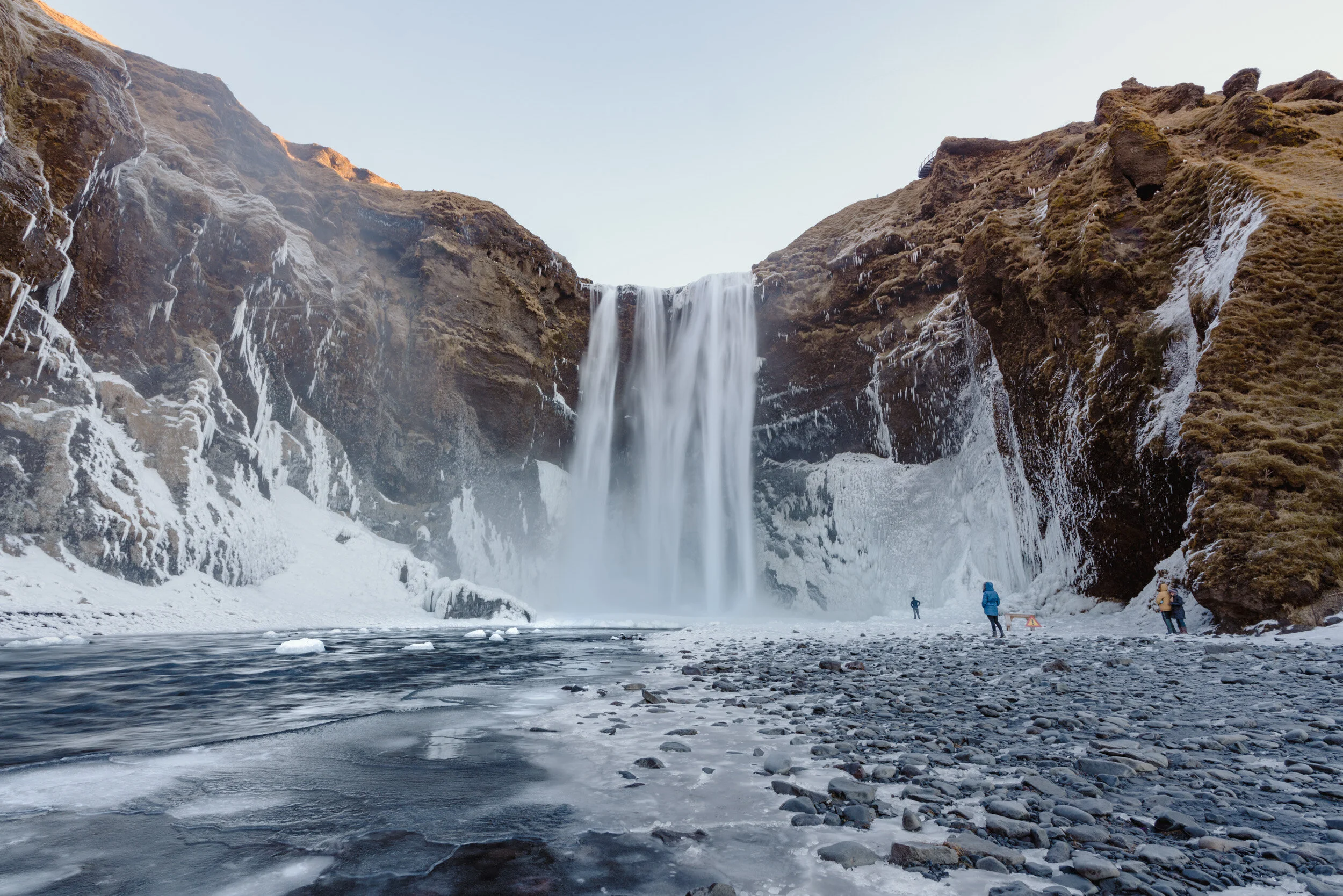 A waterfall cascading down a rocky cliff into a frozen river below. There are snow and ice on the rocks and cliff, with a few people dressed in winter clothing standing near the river's edge, observing the scene.