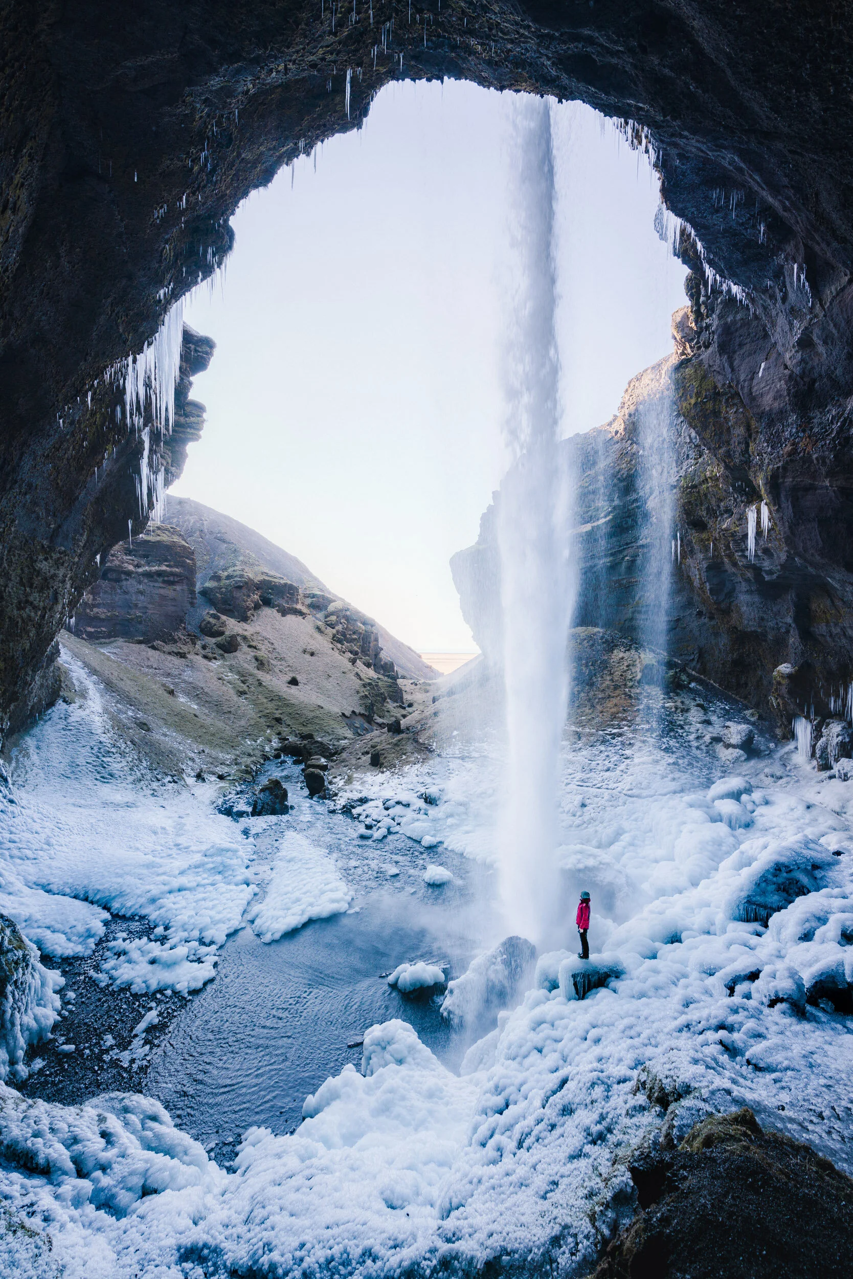 A person stands on icy rocks at the base of a large frozen waterfall inside a dark cave. The waterfall is partially frozen with icicles hanging from the cave ceiling and snow covers the ground.