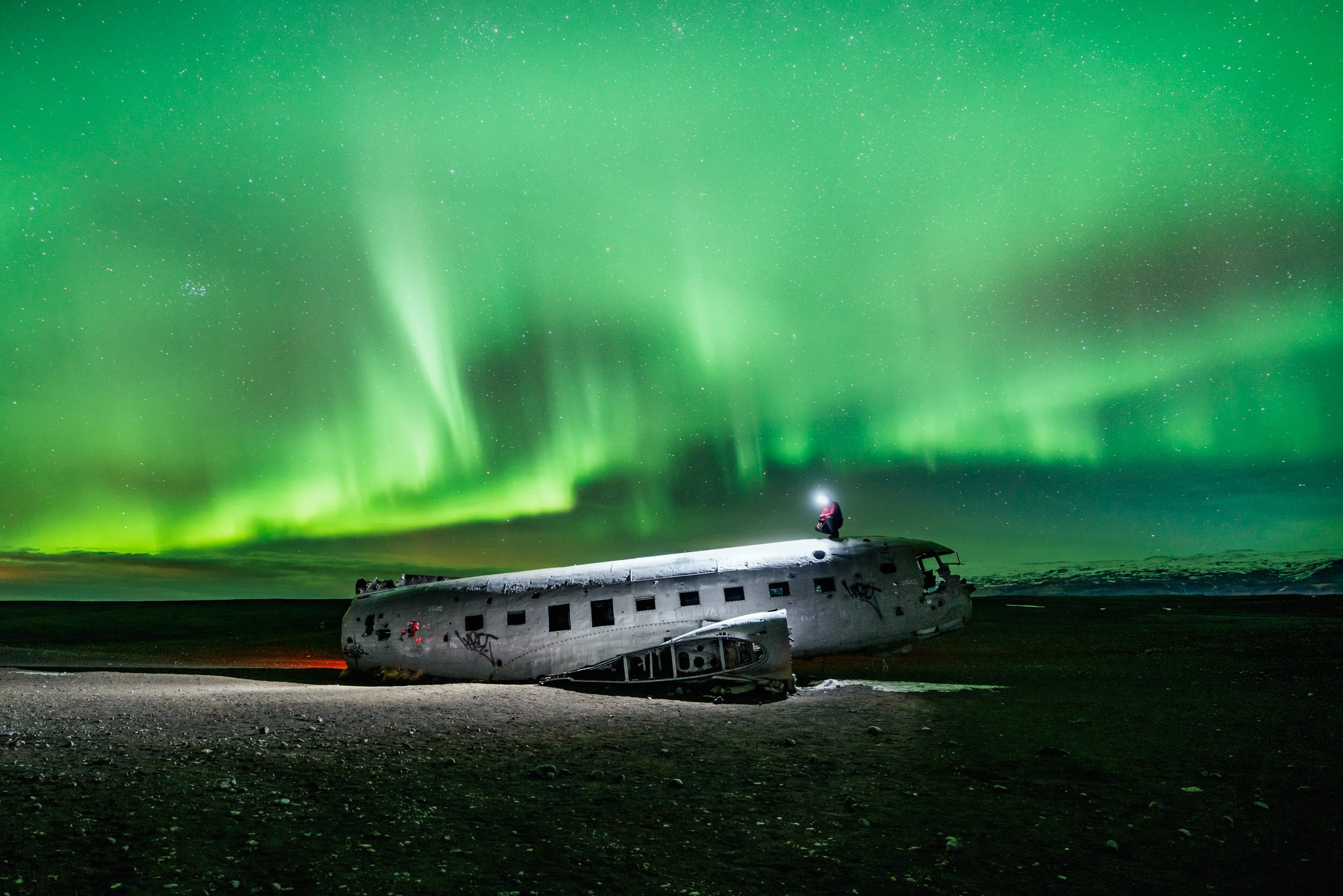 Northern lights illuminate a night sky above a wrecked airplane on an icy landscape, with a person kneeling on the plane.