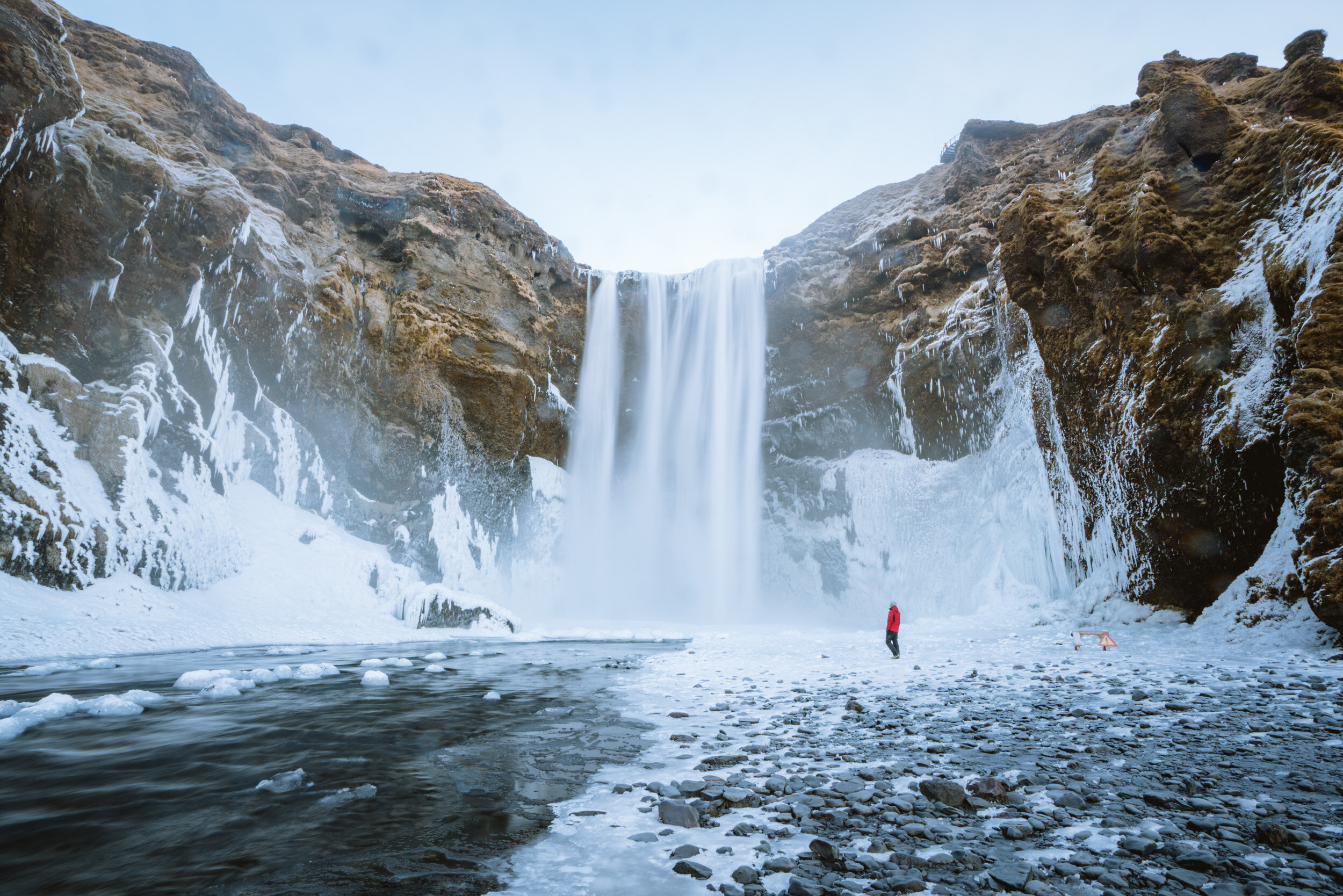 A person in a red jacket standing by a partially frozen river beneath a tall, icy waterfall flowing over rocky cliffs on a cold winter day.