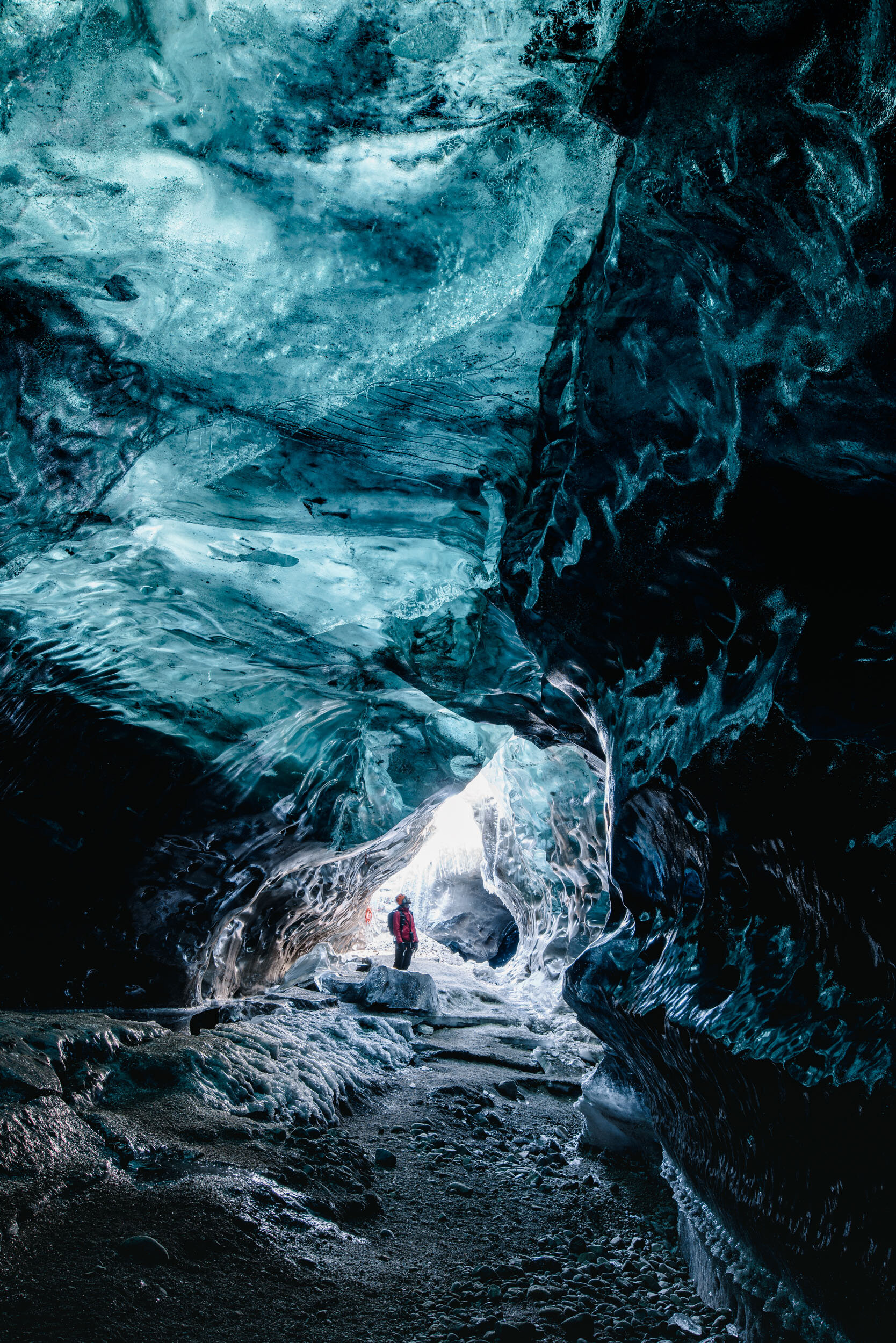 A person in a red jacket standing inside an icy, cavernous ice tunnel with smooth, glossy, blue and black ice walls.
