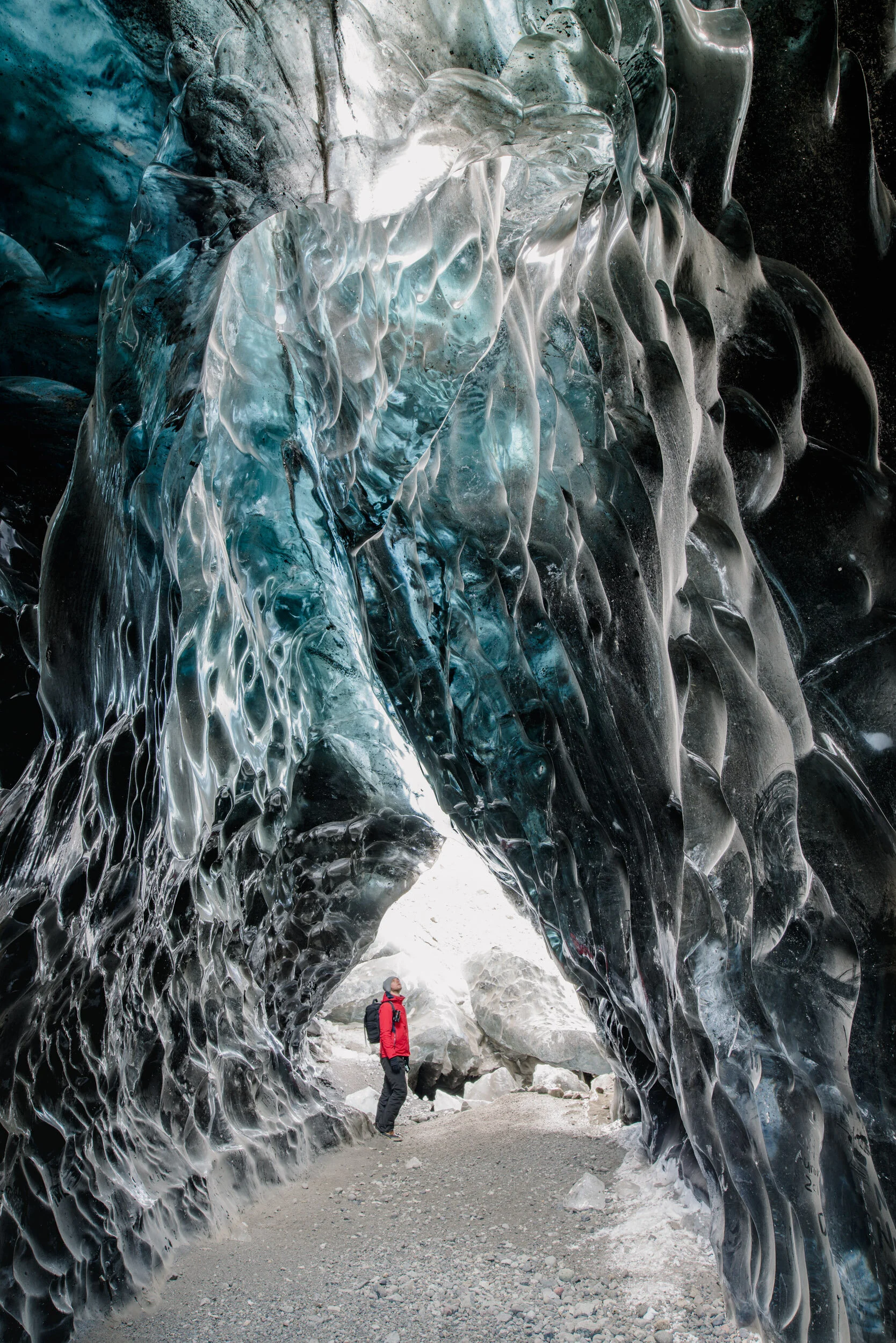 A person standing inside an ice cave with intricate ice formations on the walls and ceiling, wearing a red jacket and black pants.