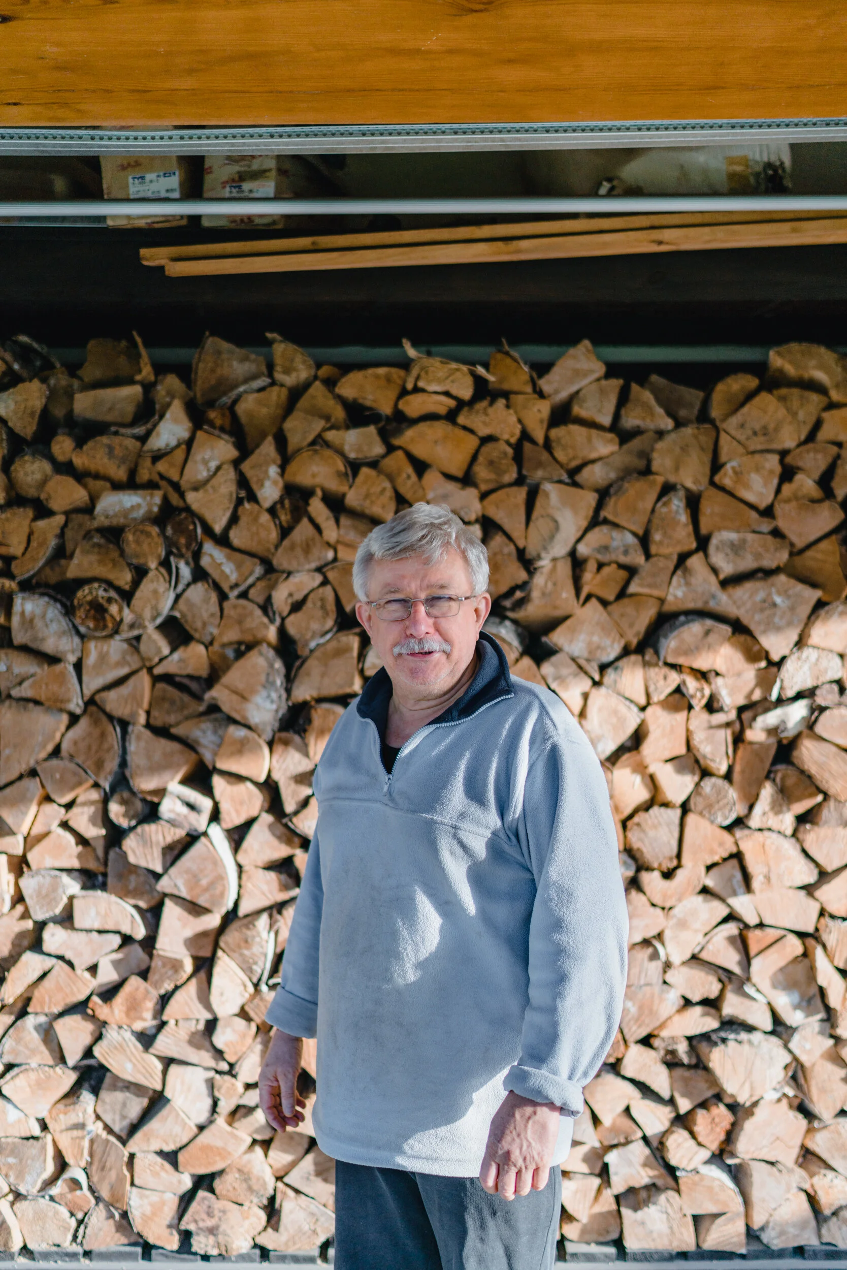 An older man with gray hair and glasses smiling while standing in front of a large stack of firewood.