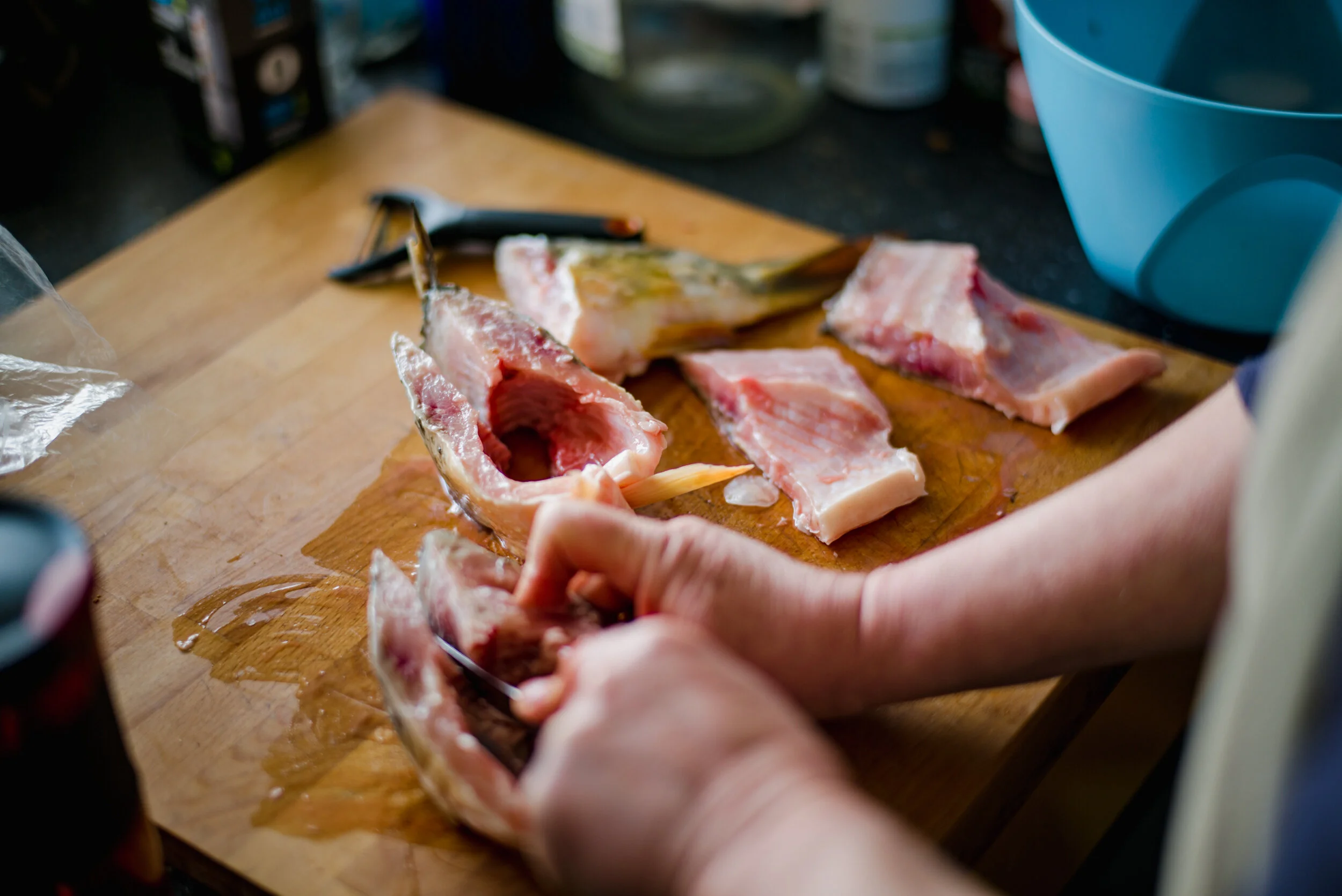 Person cutting raw pork on a wooden cutting board in a kitchen. Blue bowl nearby, some tools and ingredients visible in the background.